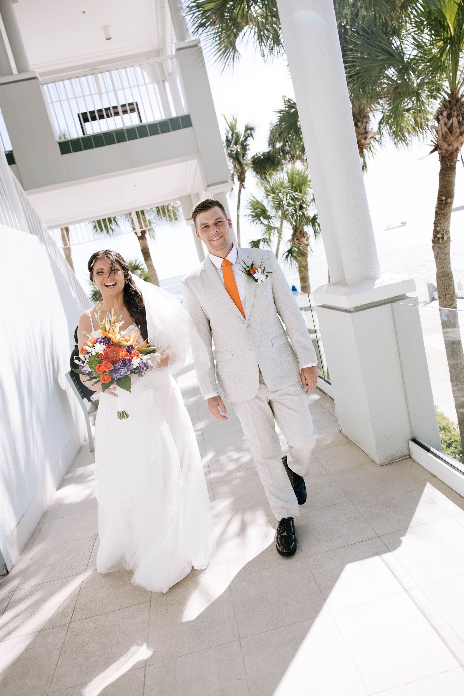 Bride and groom walking together outdoors, groom in white suit with orange tie.