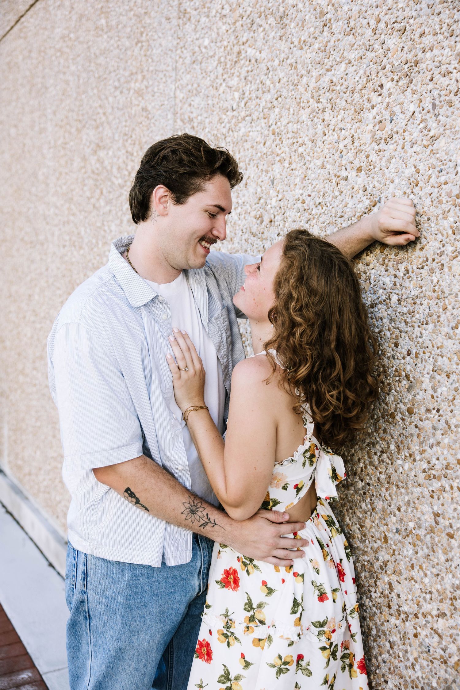 Smiling couple sharing a romantic moment against a textured wall, man with tattoos in blue shirt, woman in floral dress.