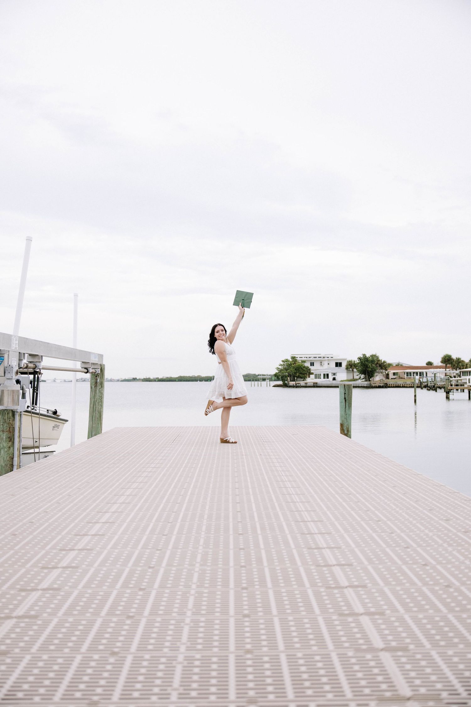 Woman in white dress celebrating on a waterfront dock, joyfully raising a book while balancing on one leg.Photographers at Anna Maria Island
