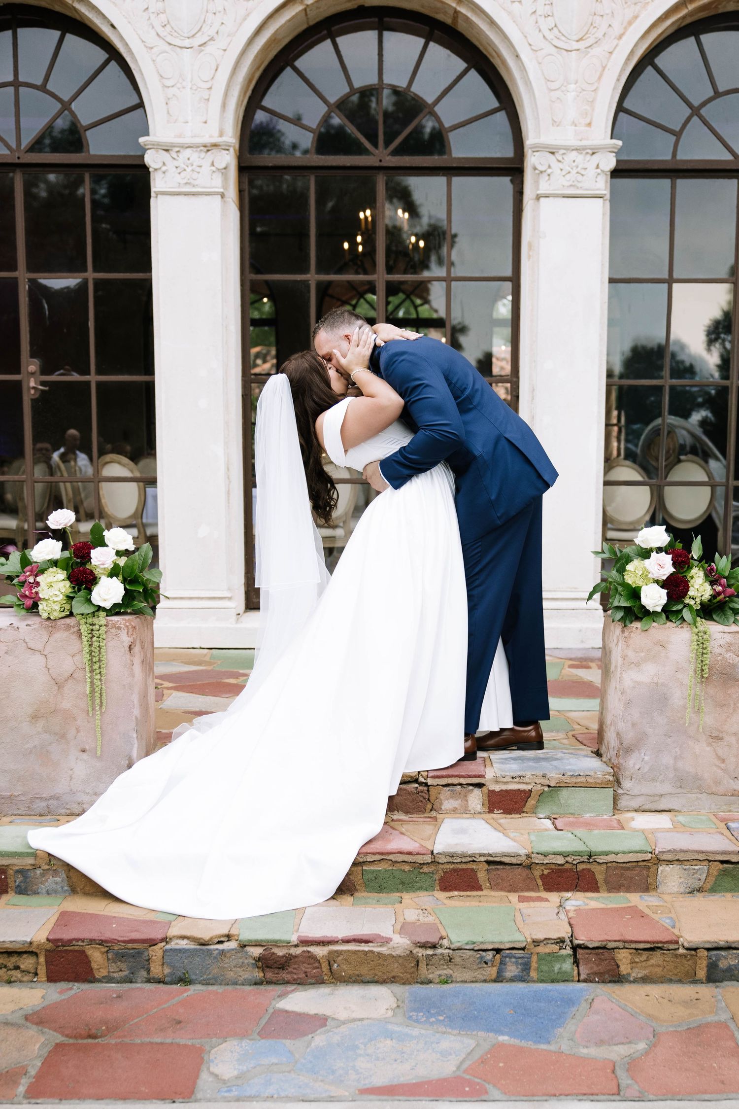 Bride and groom share a romantic dip kiss on colorful stone steps outside an elegant venue with arched windows.