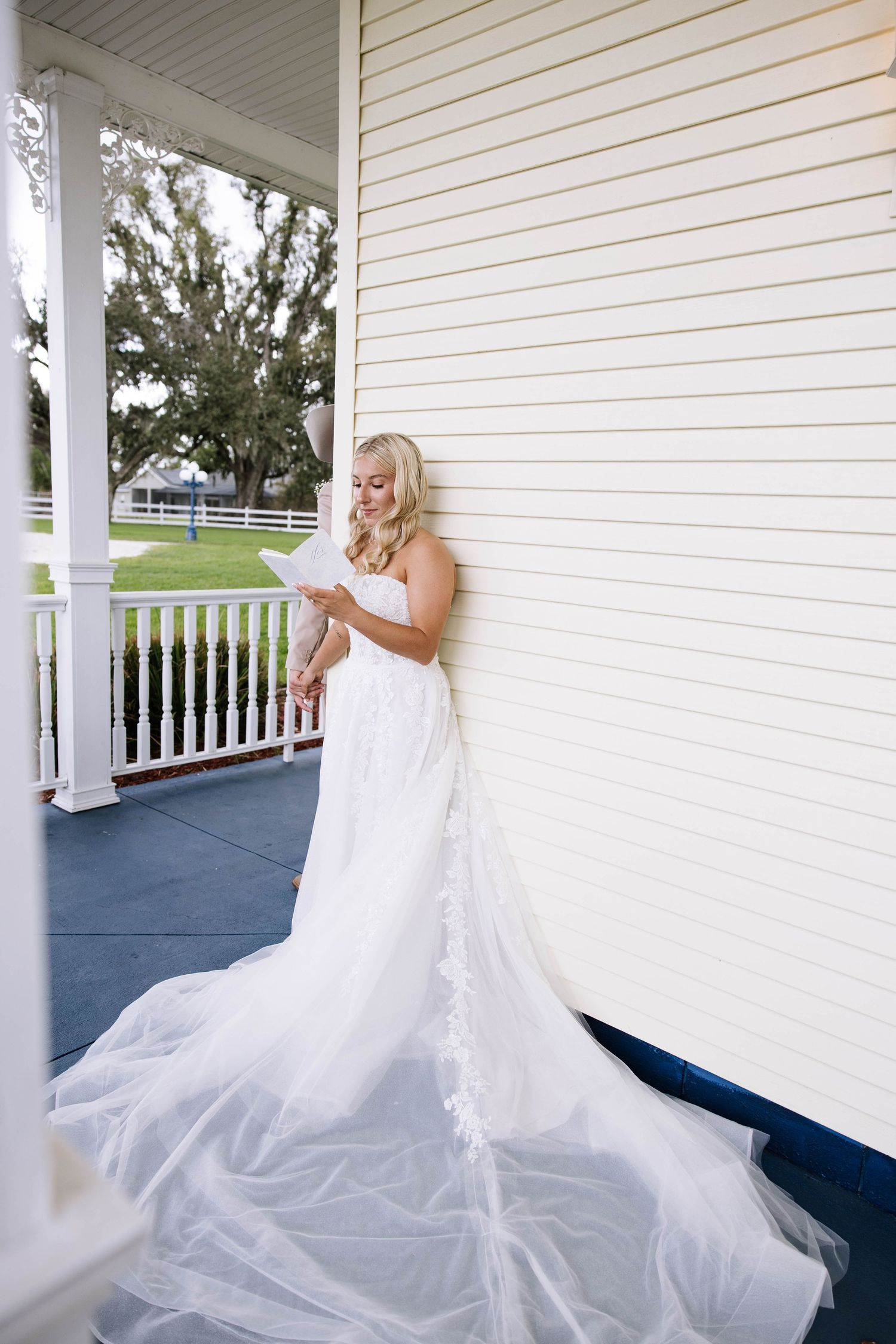 Blonde bride in strapless white gown with cathedral train reading vows on a porch.