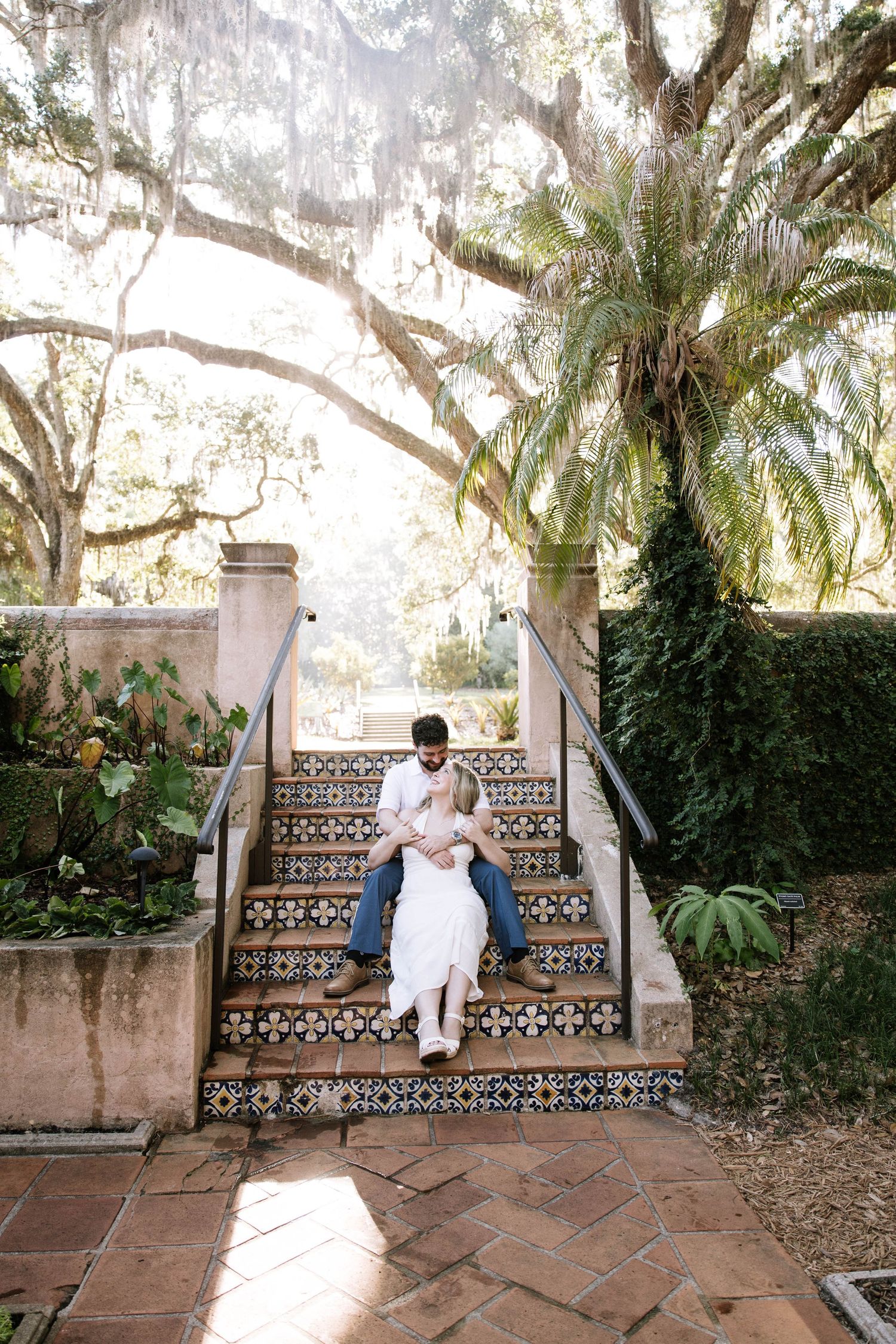 Couple sitting closely together on decorative tiled steps surrounded by lush trees and tropical garden landscape.