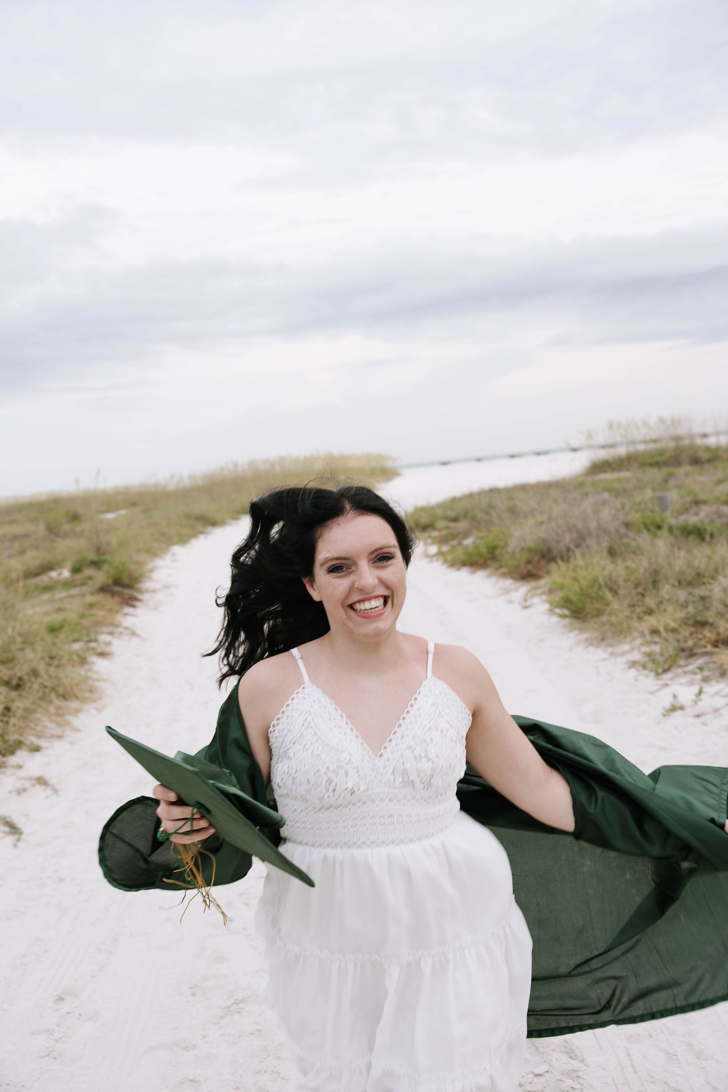 Smiling woman in white dress holding a flowing green fabric on a sandy beach path on a cloudy day. Photographers at Anna Maria Island FL.