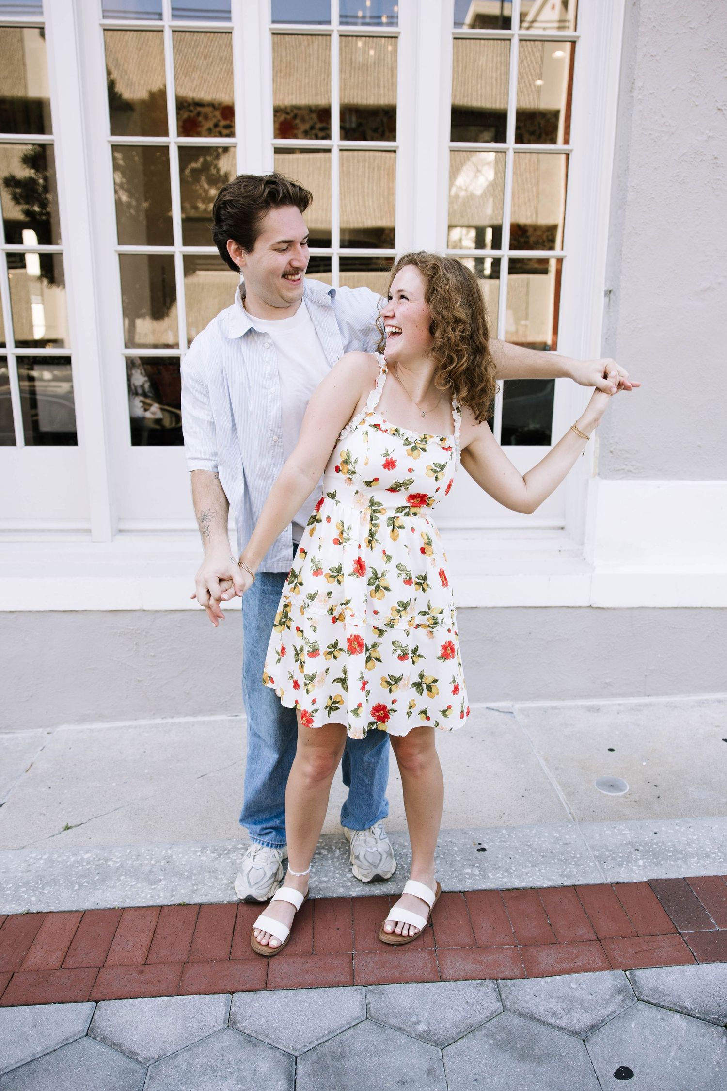A joyful couple holds hands and laughs together on a sidewalk, woman wearing a floral sundress.