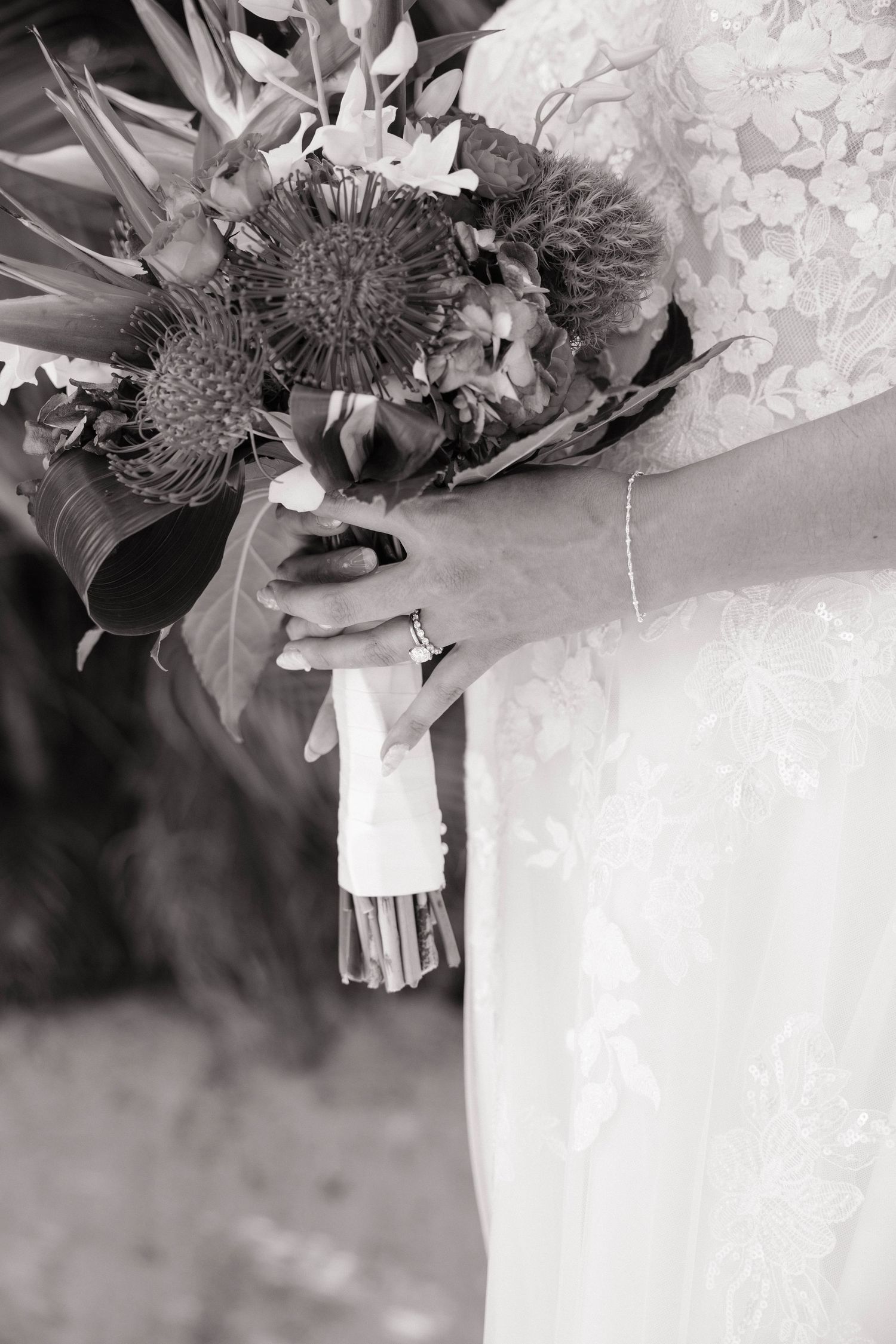 A black and white close-up of a bride holding a tropical floral bouquet wrapped in white ribbon.