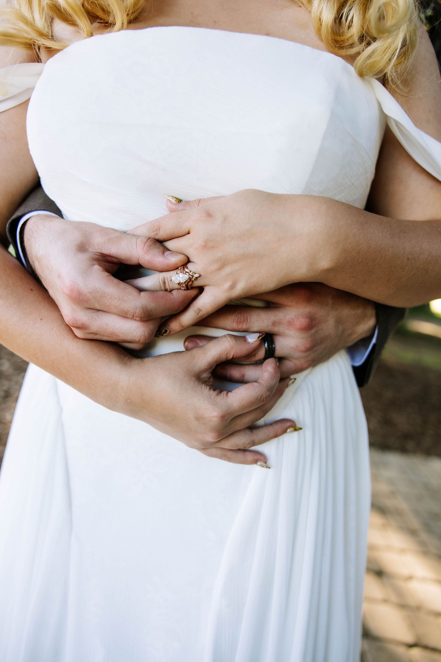 A groom embraces a bride in a white dress, their hands intertwined showing a gold diamond engagement ring.