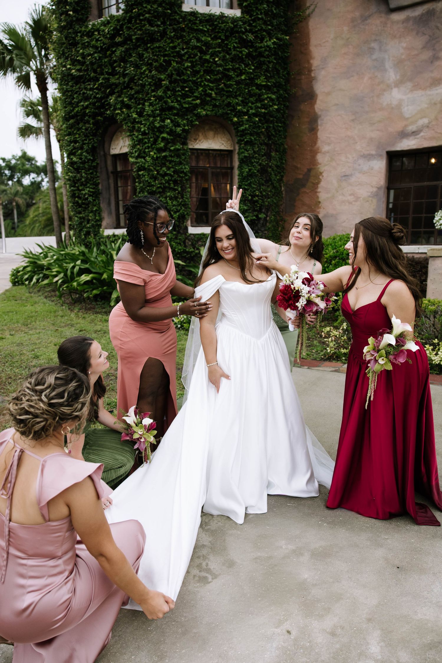 Bride in white gown surrounded by bridesmaids in pink and red dresses outside ivy-covered stone building.
