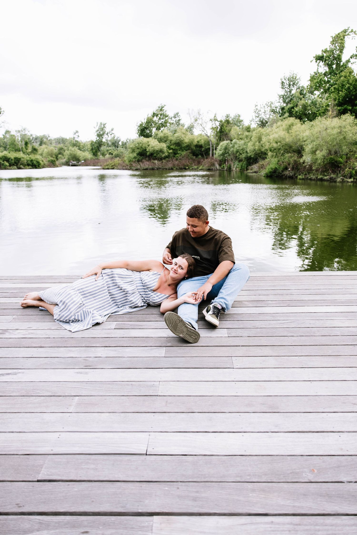Couple relaxing on a wooden dock by a calm lake, woman lying in striped dress with head on mans lap.