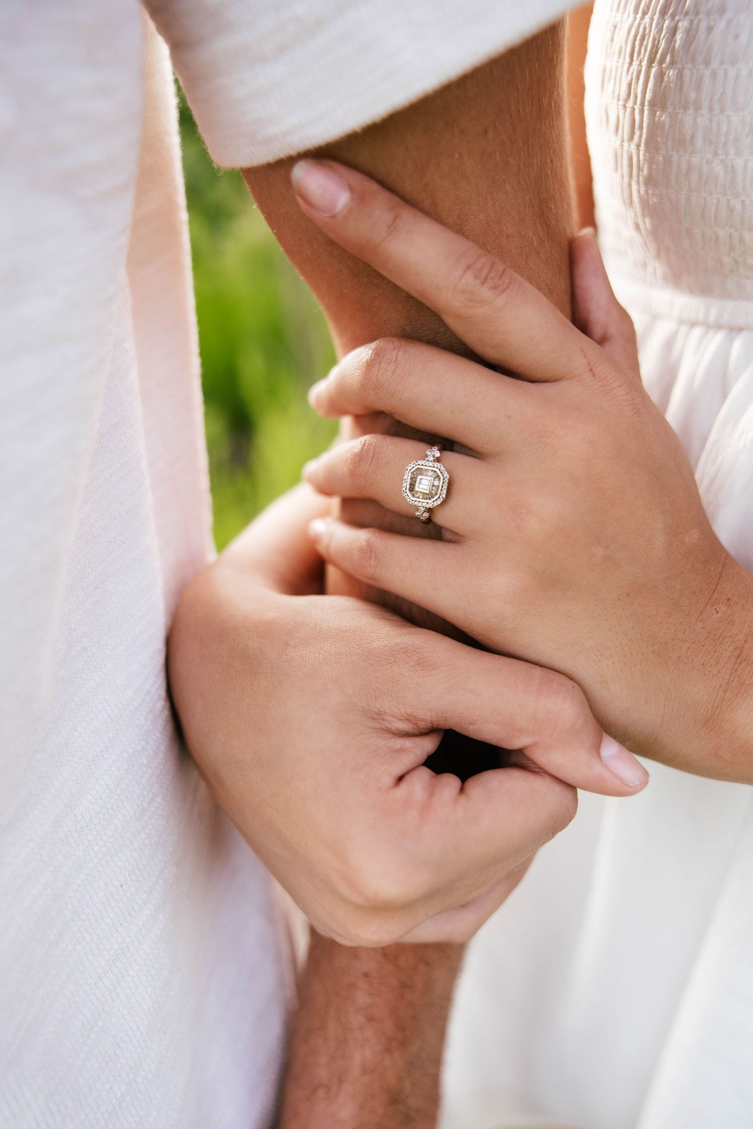 Close-up of a woman's hand wearing a stunning diamond halo engagement ring while holding hands with her partner outdoors.