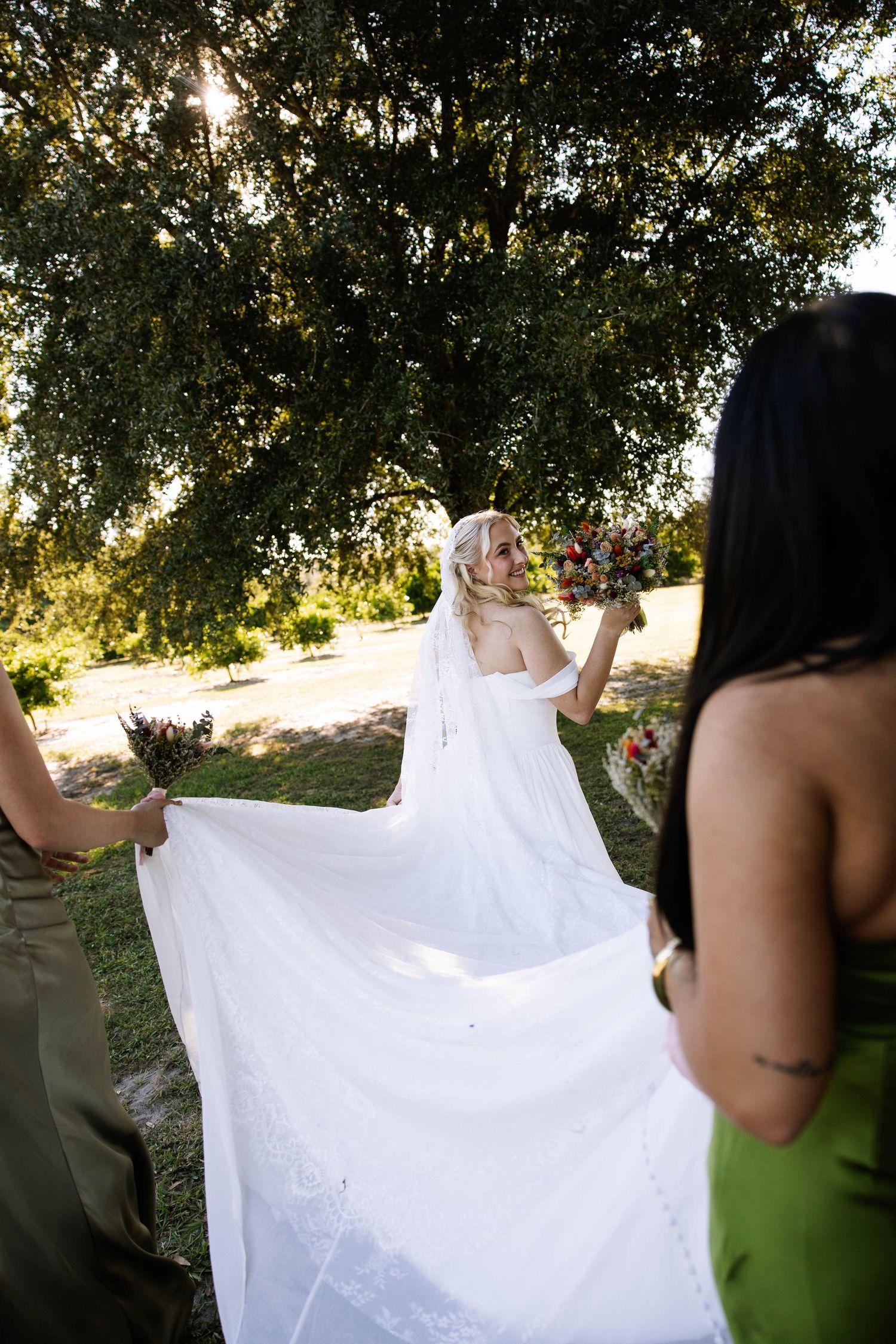 Bride in white gown and veil walks outdoors as bridesmaids in green dresses carry her train beneath a large tree.