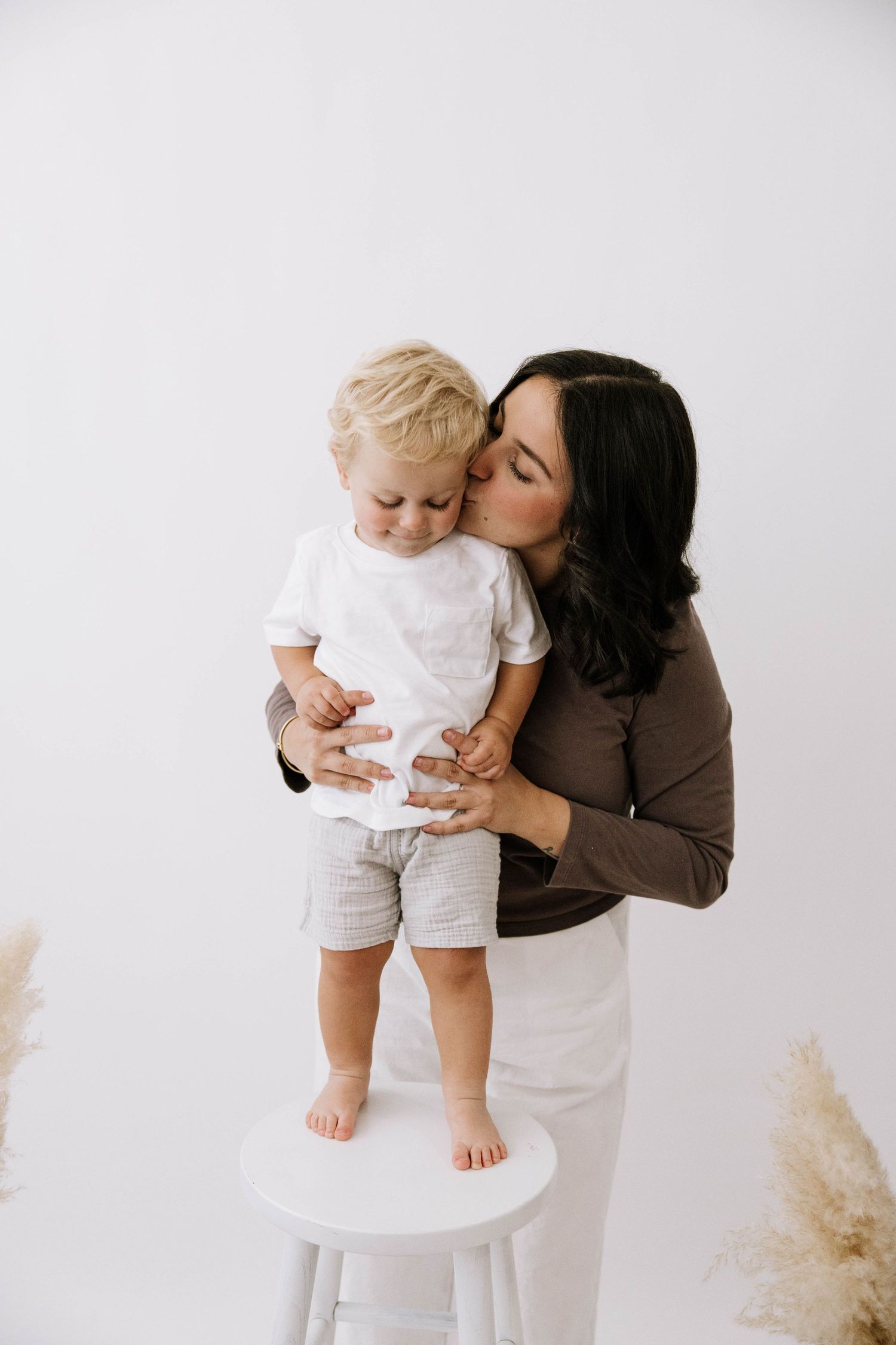 Dark-haired mother in brown top kissing her blonde toddler standing on white stool in bright studio setting.Photography Studio in Bartow