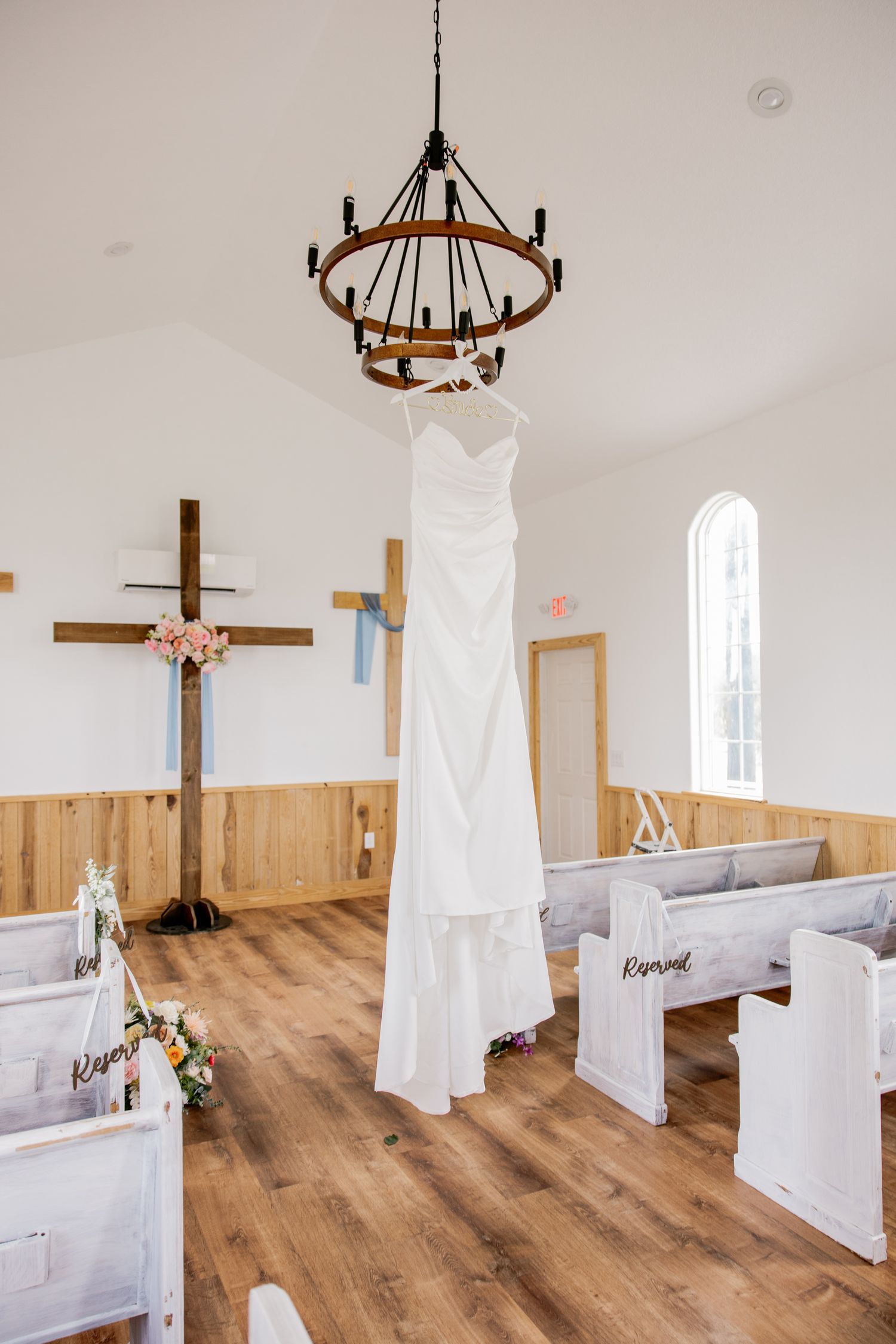 White wedding gown hanging from rustic chandelier in charming small chapel with wooden pews and crosses.