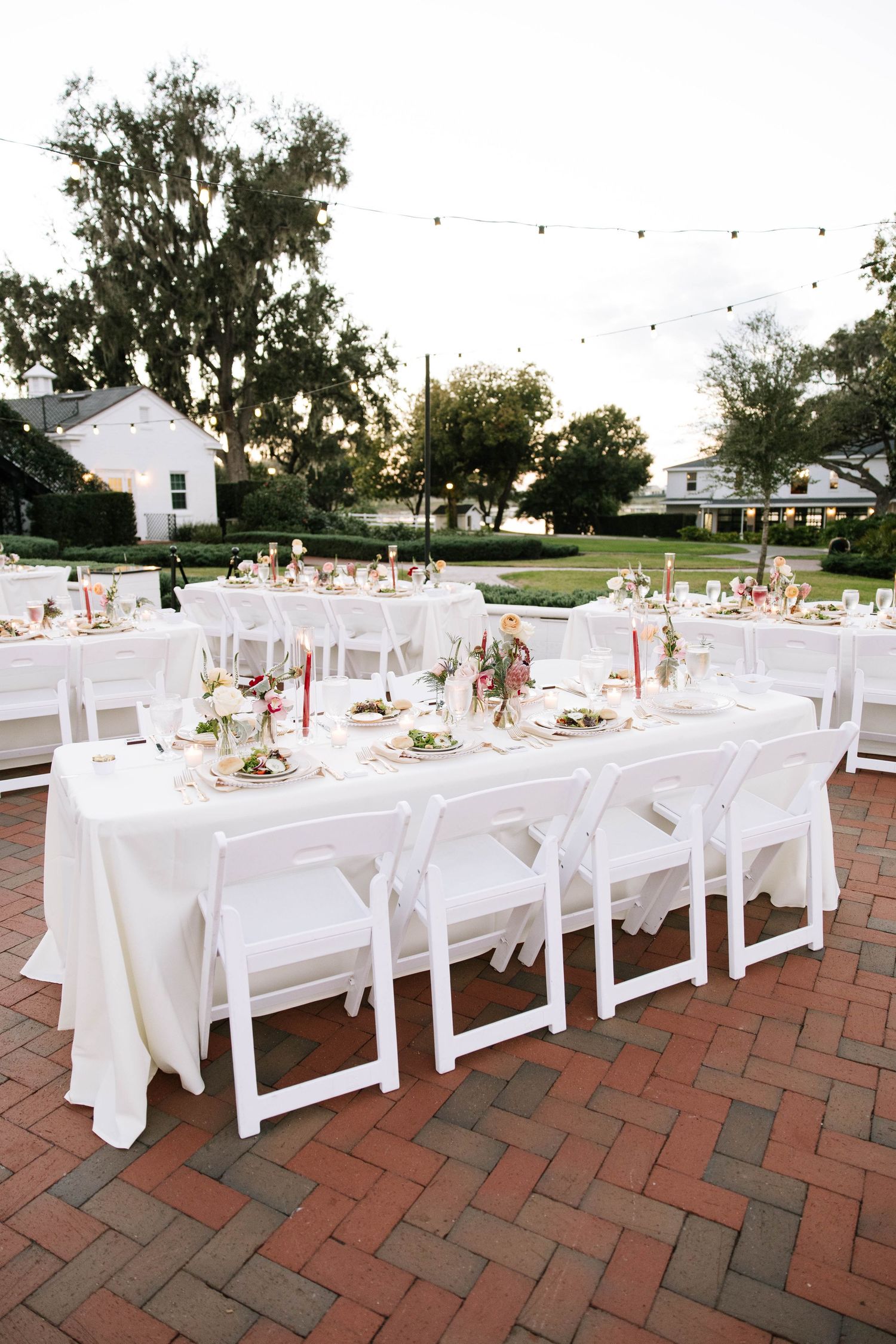 Elegant outdoor wedding reception tables with white linens, floral centerpieces, and string lights at dusk.