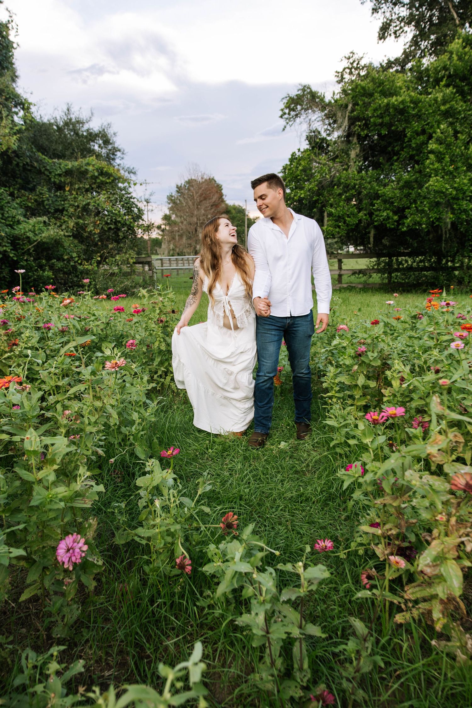 A couple walks hand-in-hand through a lush flower garden, smiling at each other in a romantic outdoor setting.