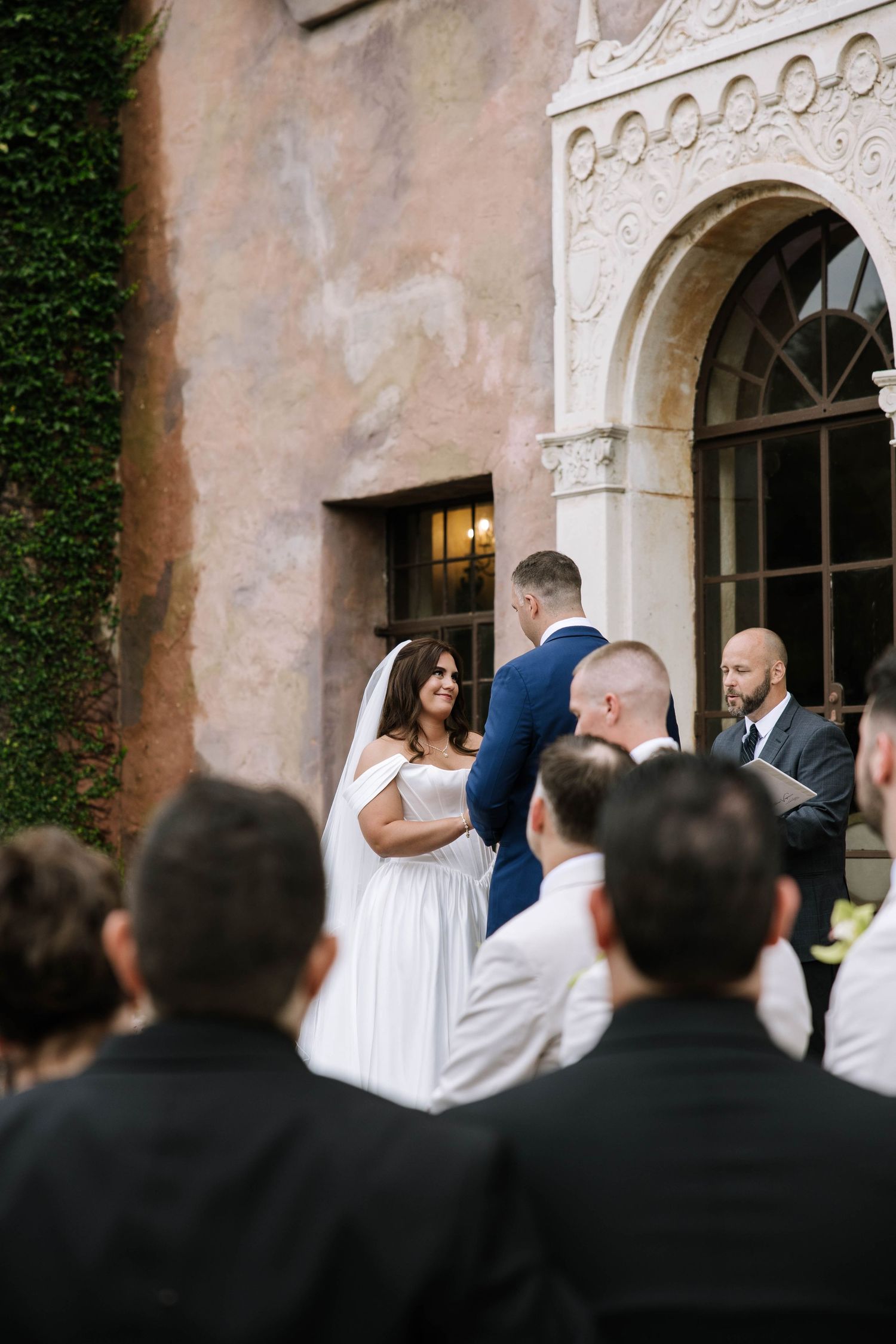 Bride and groom exchange vows outdoors at elegant venue with ivy-covered walls, officiant and guests in foreground.