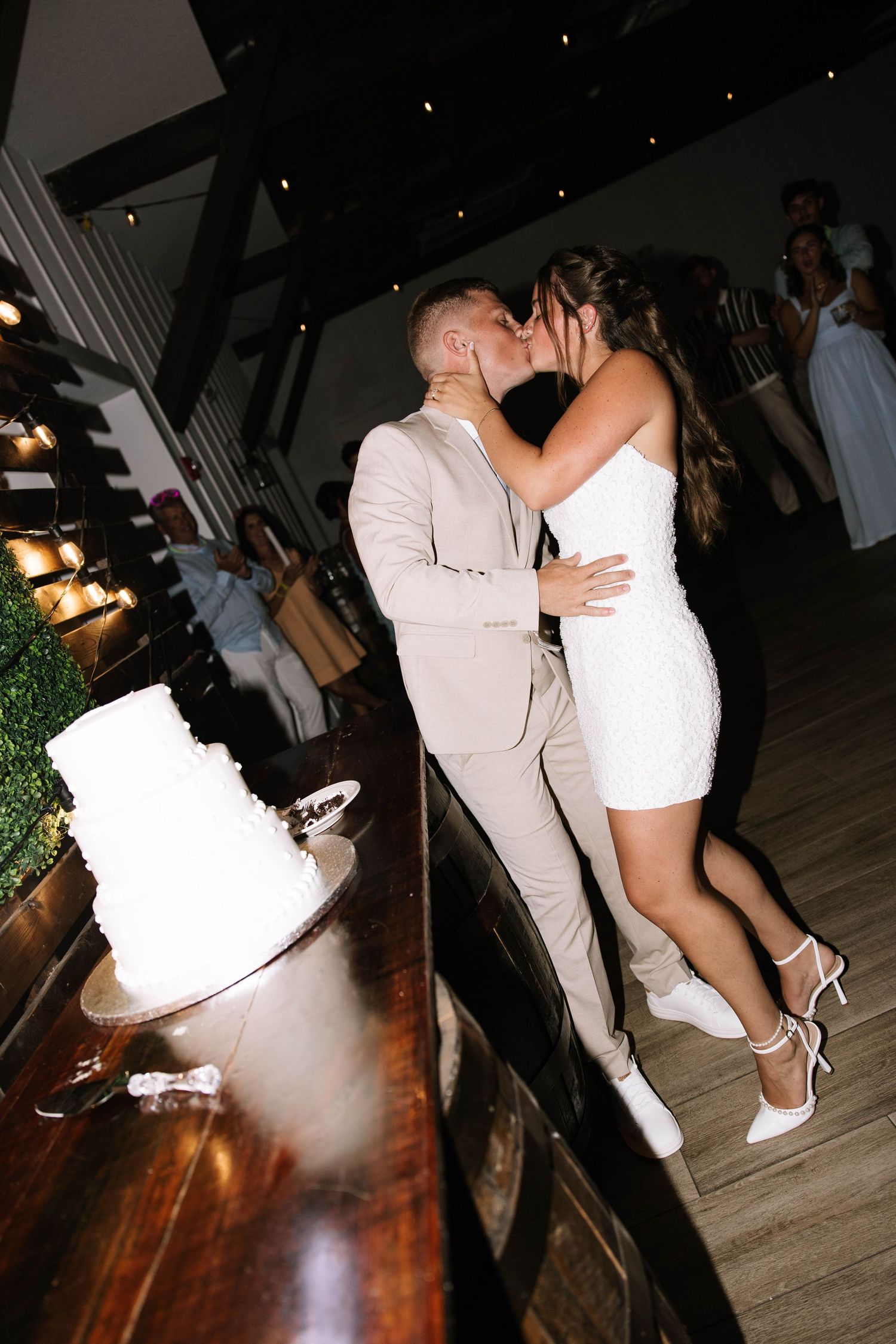 Bride and groom kiss beside white wedding cake during reception, bride wearing short white dress.