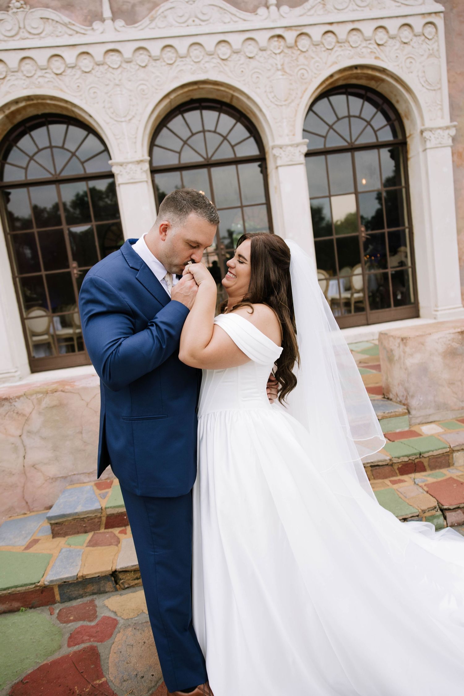 Bride and groom embracing tenderly outside elegant white venue with arched windows and colorful tile steps.