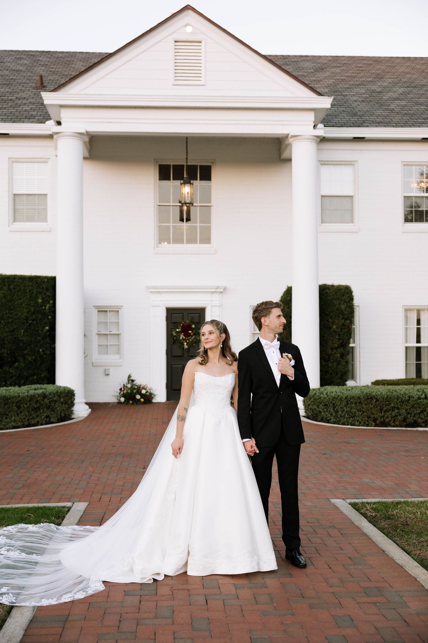 Bride in strapless ball gown with long train and groom in tuxedo pose before white colonial mansion.
