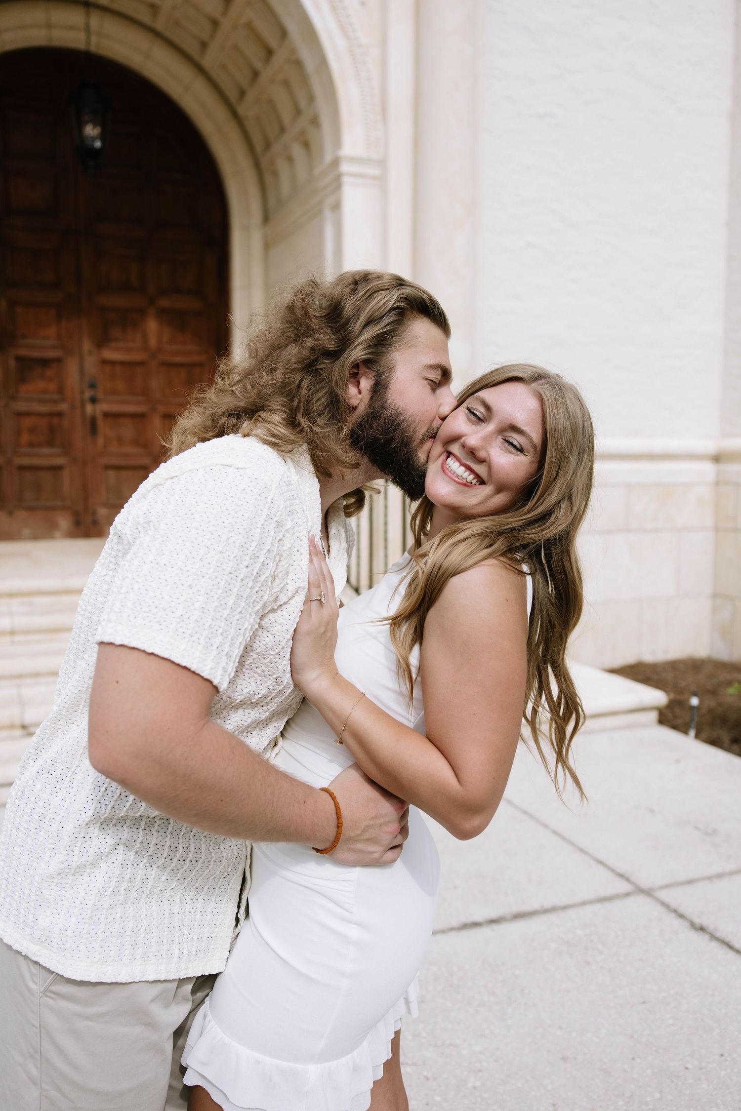 Happy couple embracing outside ornate stone building, long-haired man kissing smiling blonde woman in white dress.