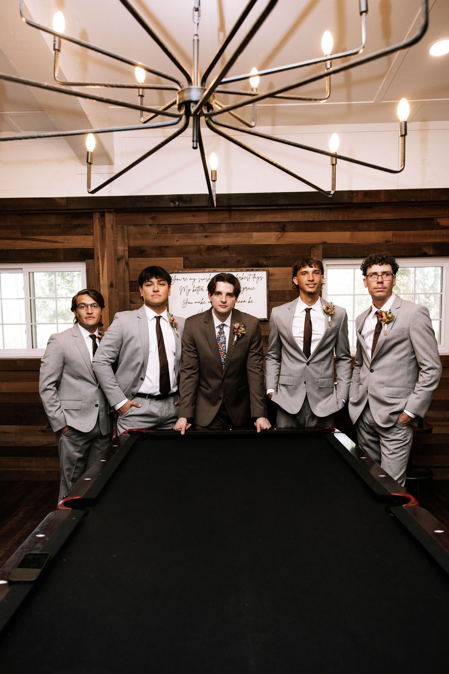 Five groomsmen in gray suits pose around a pool table under an industrial chandelier in a rustic wood-paneled room.