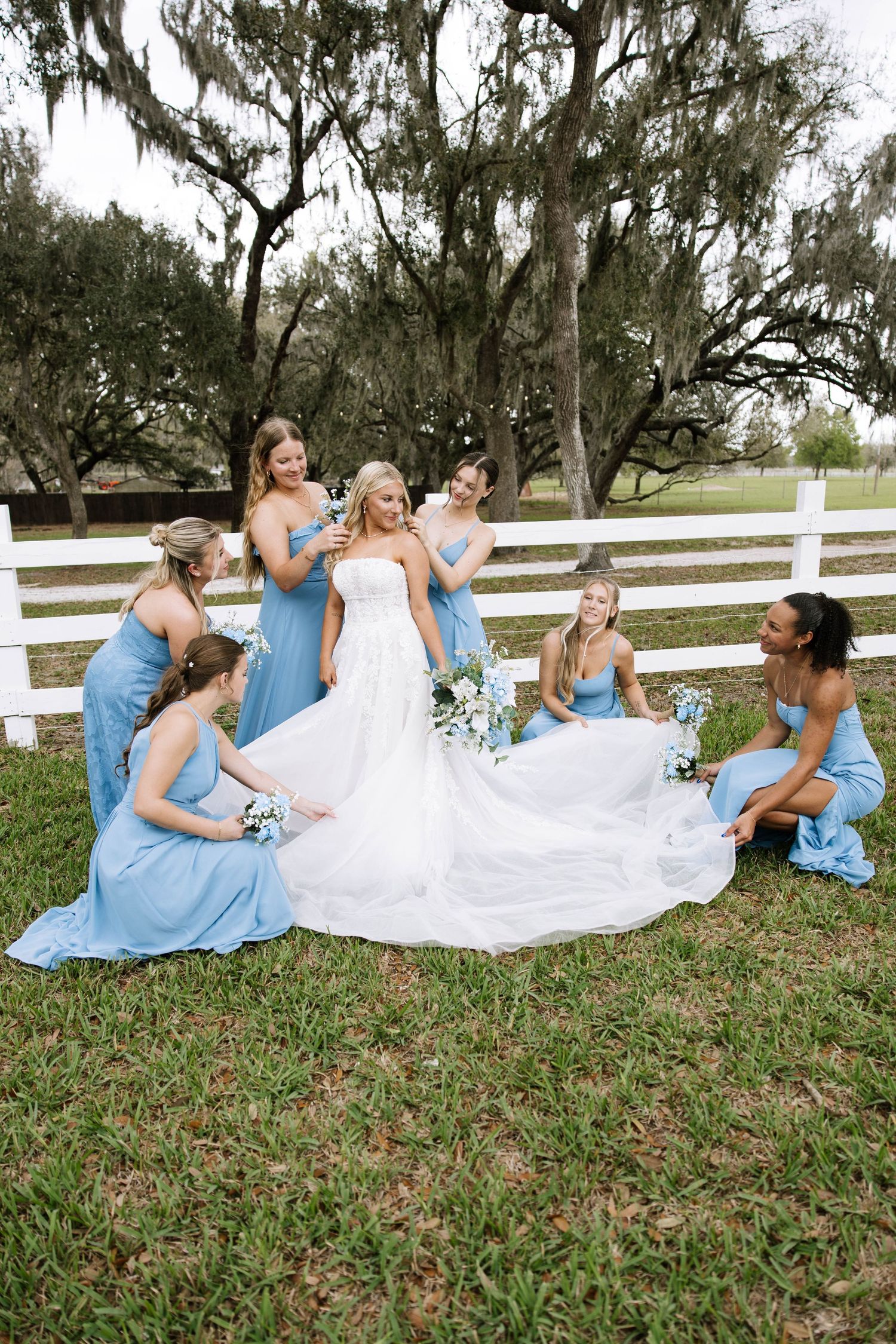 Blonde bride in white gown surrounded by bridesmaids in blue dresses outdoors.