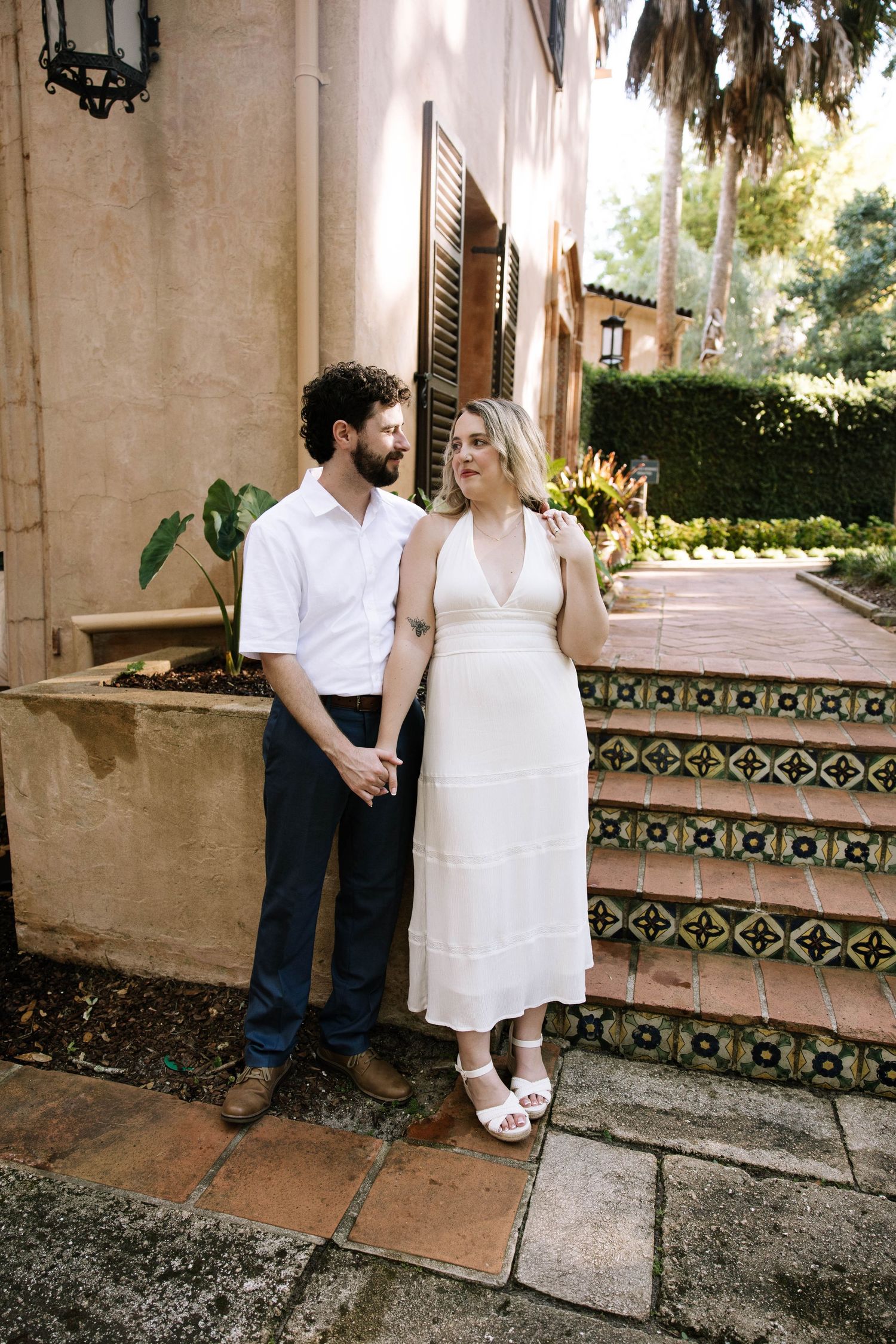 A couple holds hands beside ornate tiled steps in a lush garden, woman in elegant white midi dress.