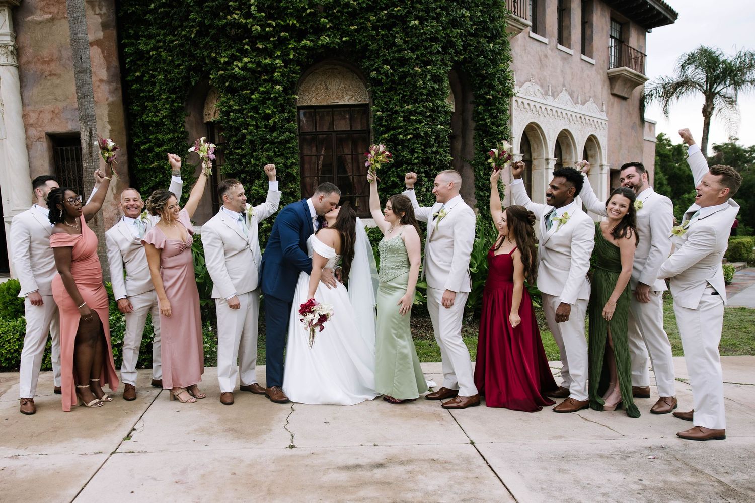 Wedding party celebrating outside ivy-covered venue, bride and groom kissing while guests cheer with raised arms.