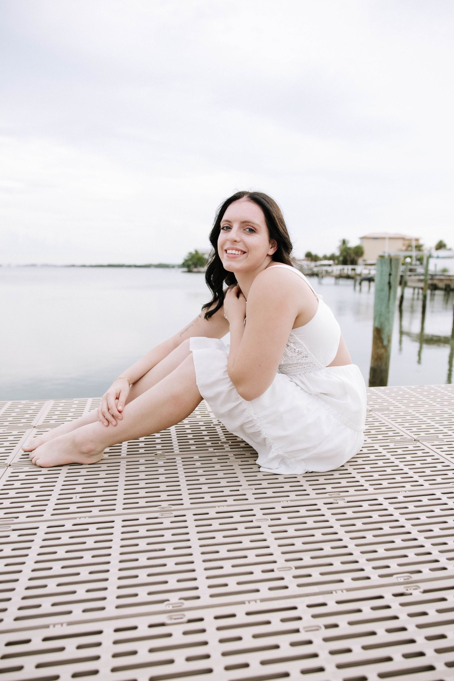 Smiling brunette woman in white dress sitting on floating dock with calm waterway and marina in background.Photographers at Anna Maria Islan