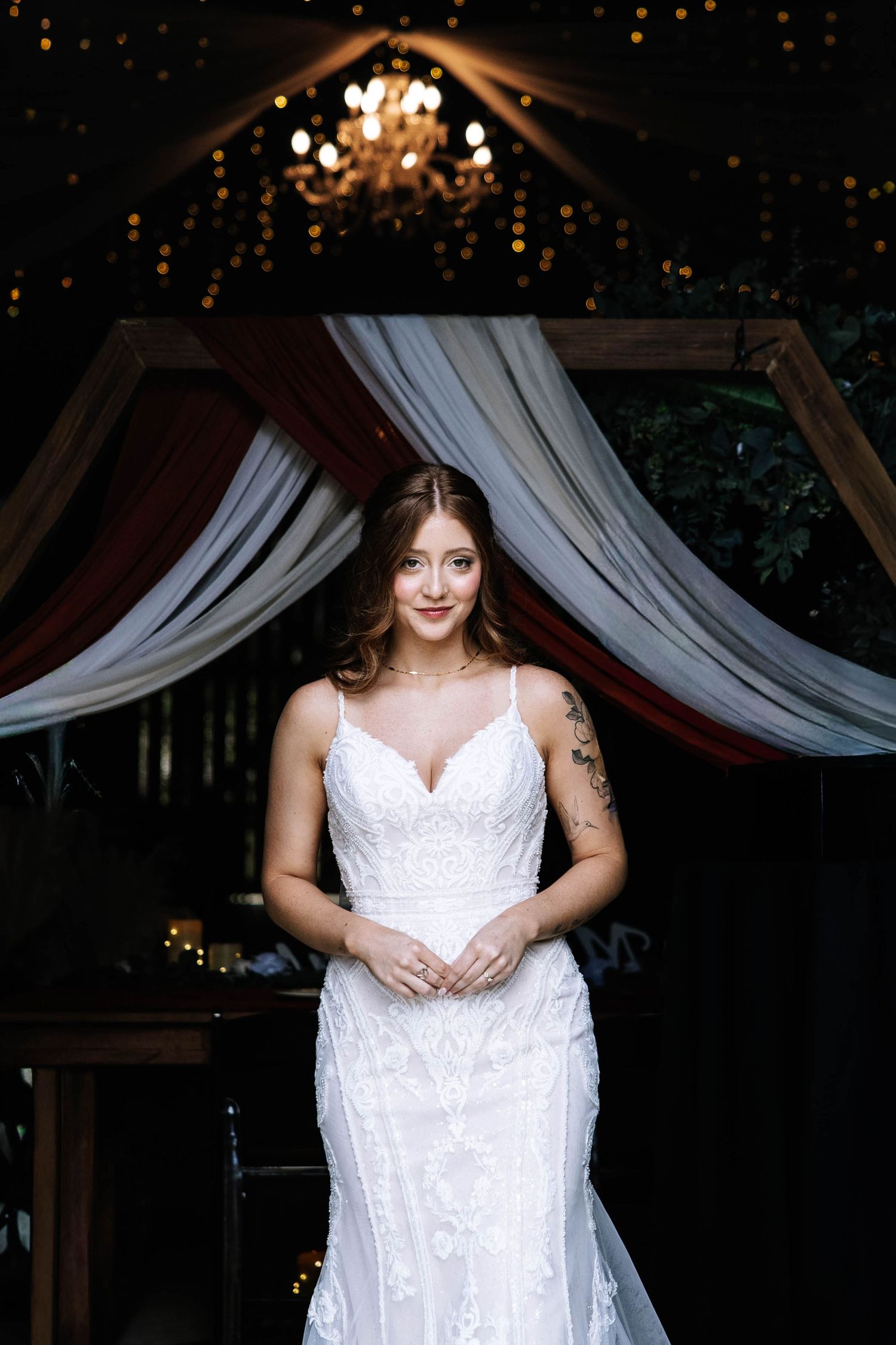 Bride in a fitted white lace wedding gown stands under a chandelier with draped fabric backdrop.