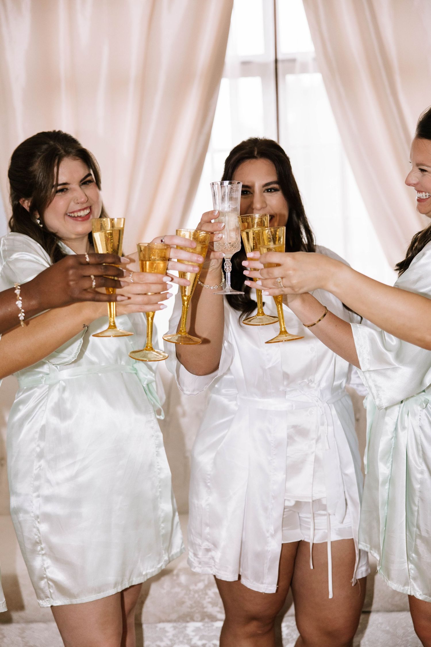 Bridesmaids in white satin robes toasting with champagne glasses during a bridal party celebration.