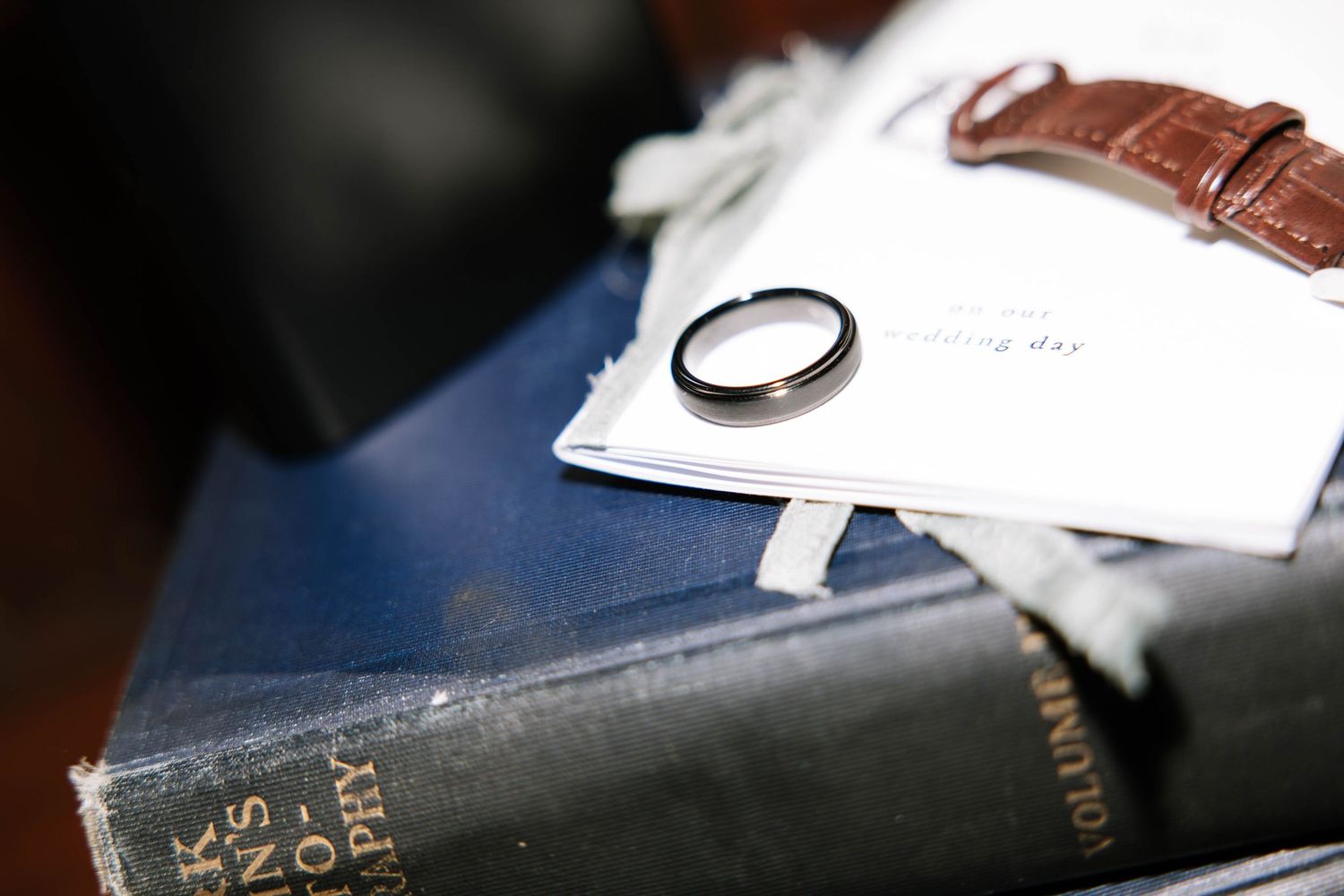 A silver wedding band resting on white papers beside a brown leather watch on top of old hardcover books.