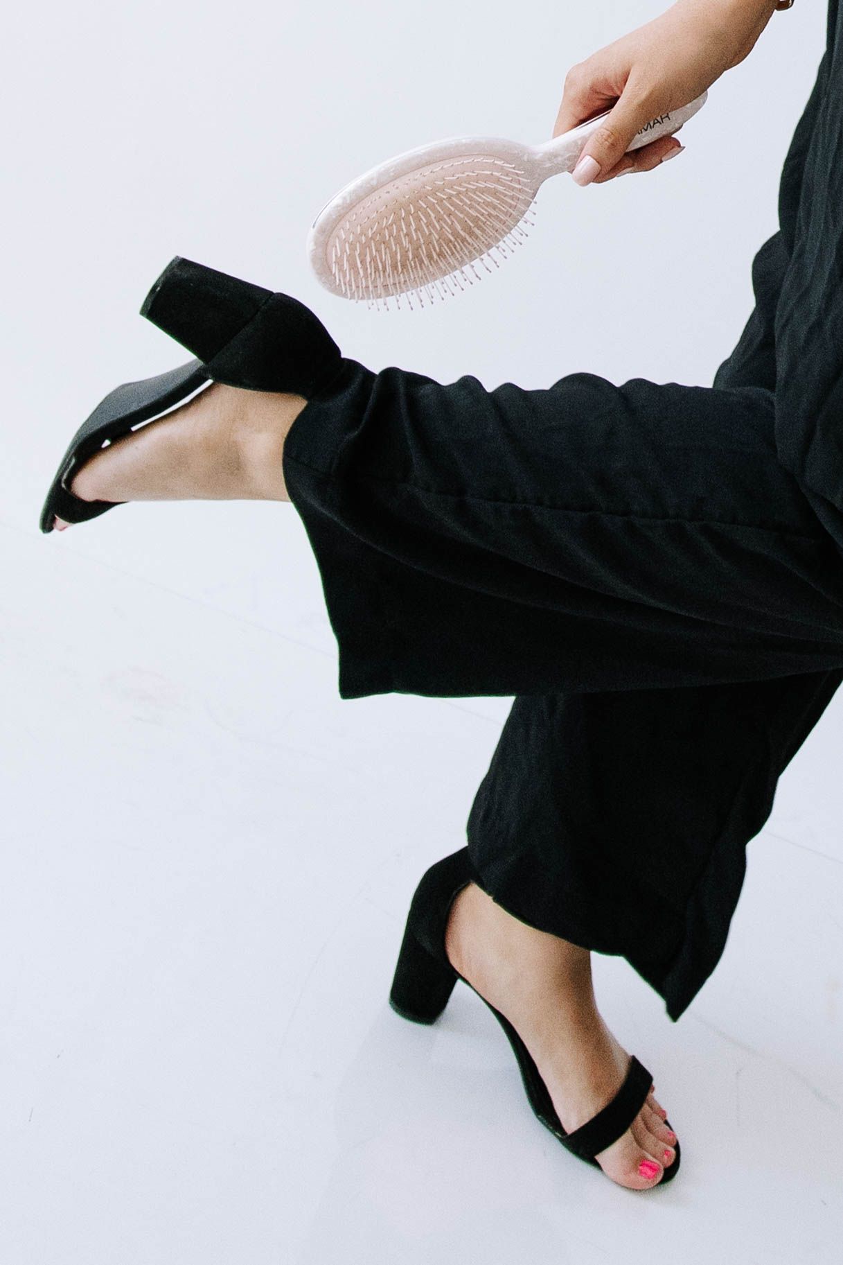 Woman in black wide-leg pants and heels holds a pink paddle hair brush against white background.Photography Studio in Bartow Florida
