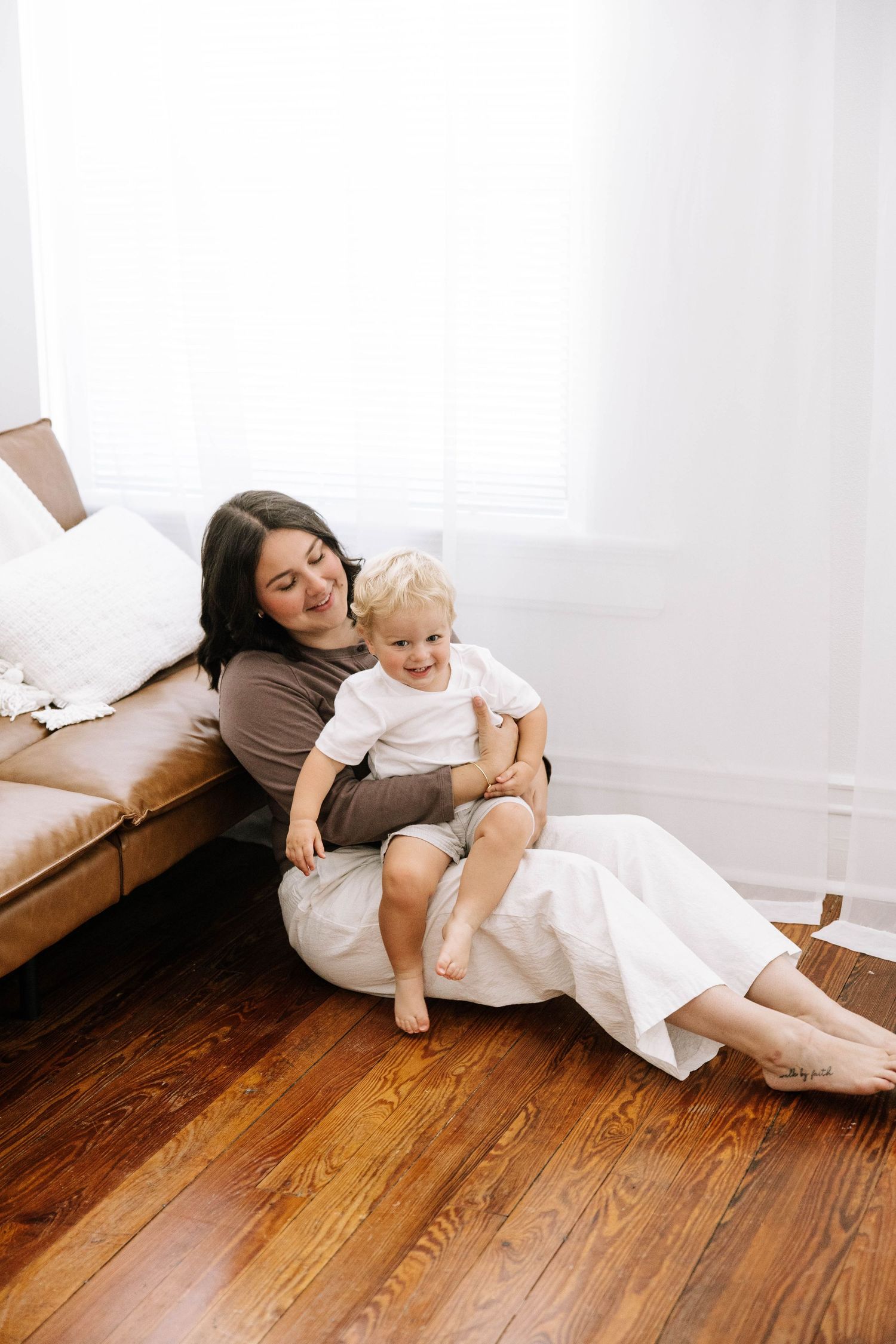 Mother in brown top sitting on hardwood floor against leather couch, smiling while holding her blonde toddler.Photography Studio in Bartow