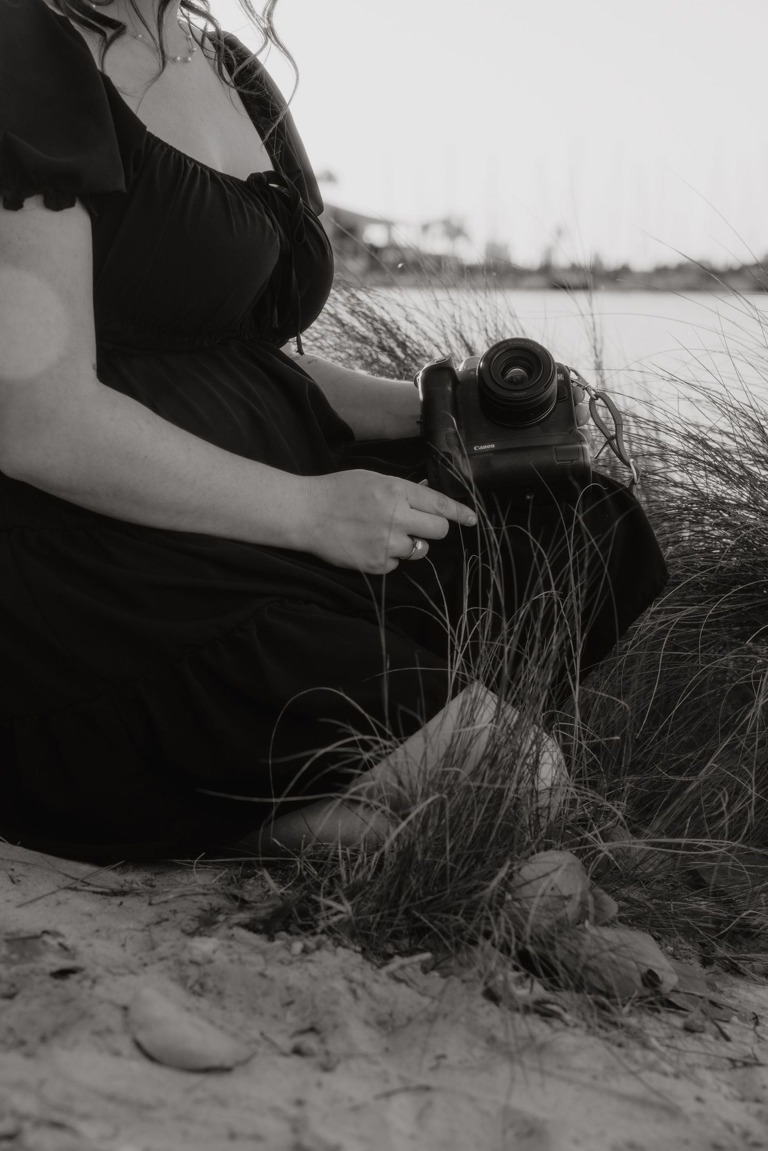 Black and white photo of a woman in a black dress sitting on sandy dunes holding a Canon camera.