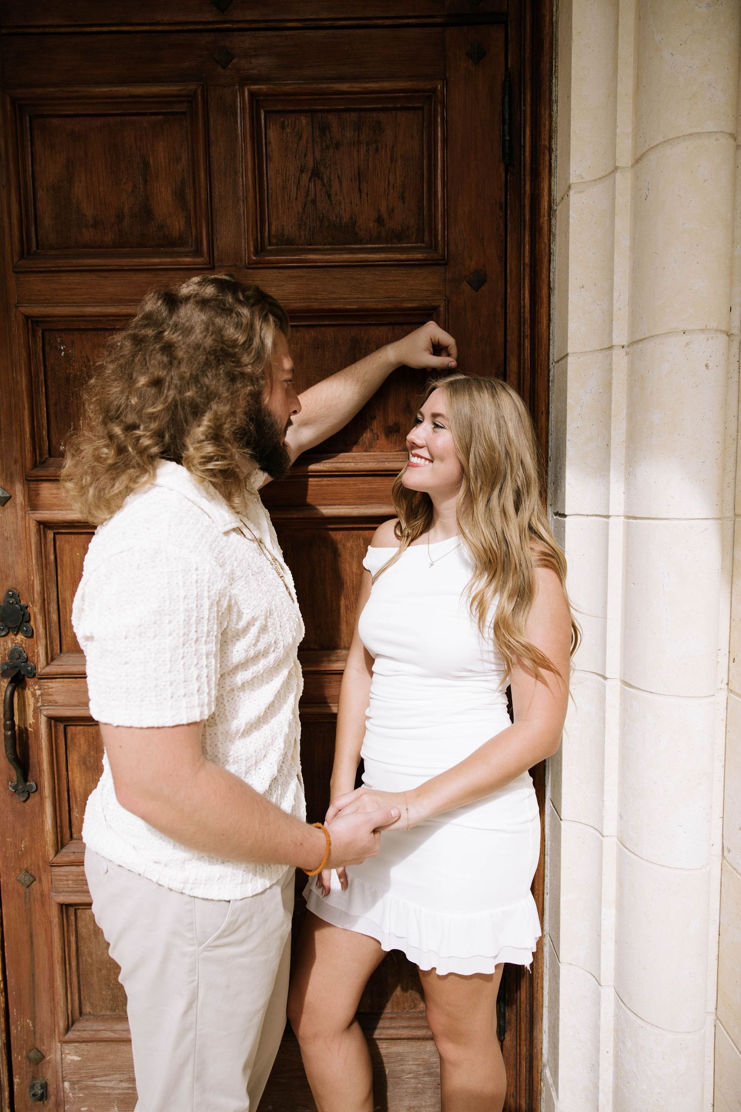 Couple in white outfits hold hands and smile by ornate wooden door.
