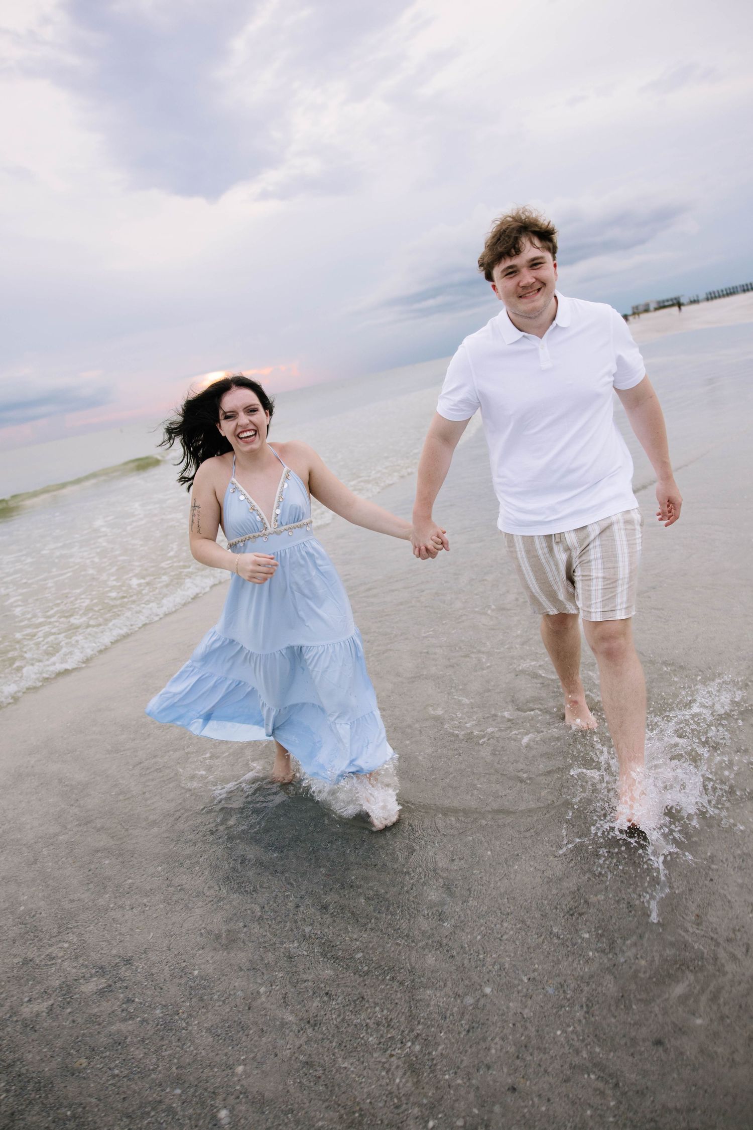 Happy couple holding hands and running through ocean waves on beach, woman in blue tiered dress and man in white polo.