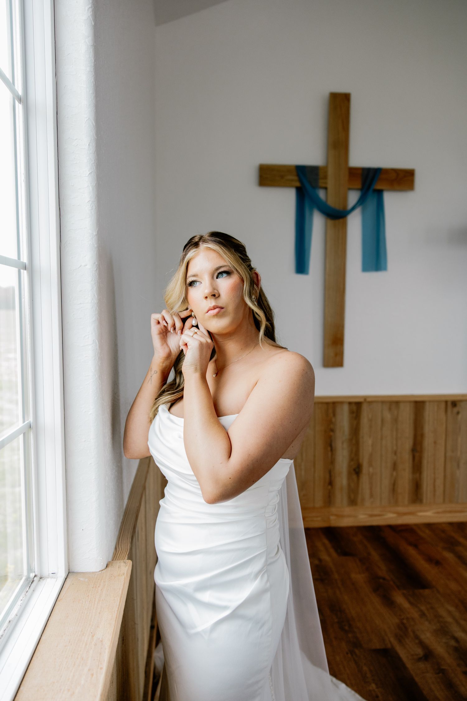 Blonde bride in white gown putting on earrings near window with wooden cross and blue ribbon on wall behind her.