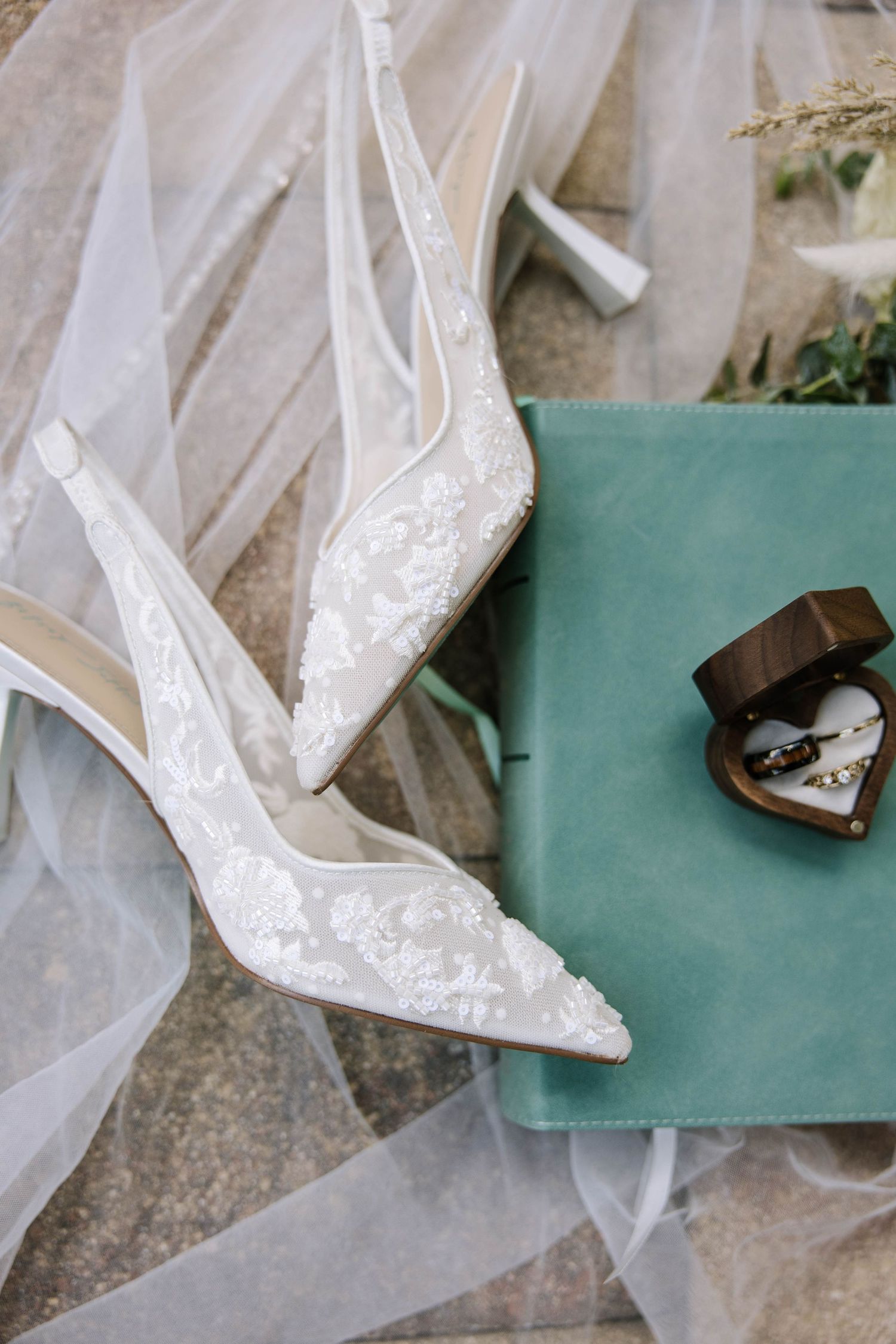White lace bridal heels resting on a veil beside a teal velvet ring box with wedding rings on stone.