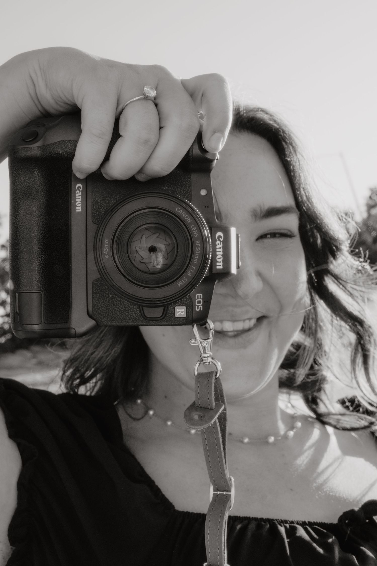 Smiling young woman holding a Canon EOS camera up to her face in a black and white portrait photo.