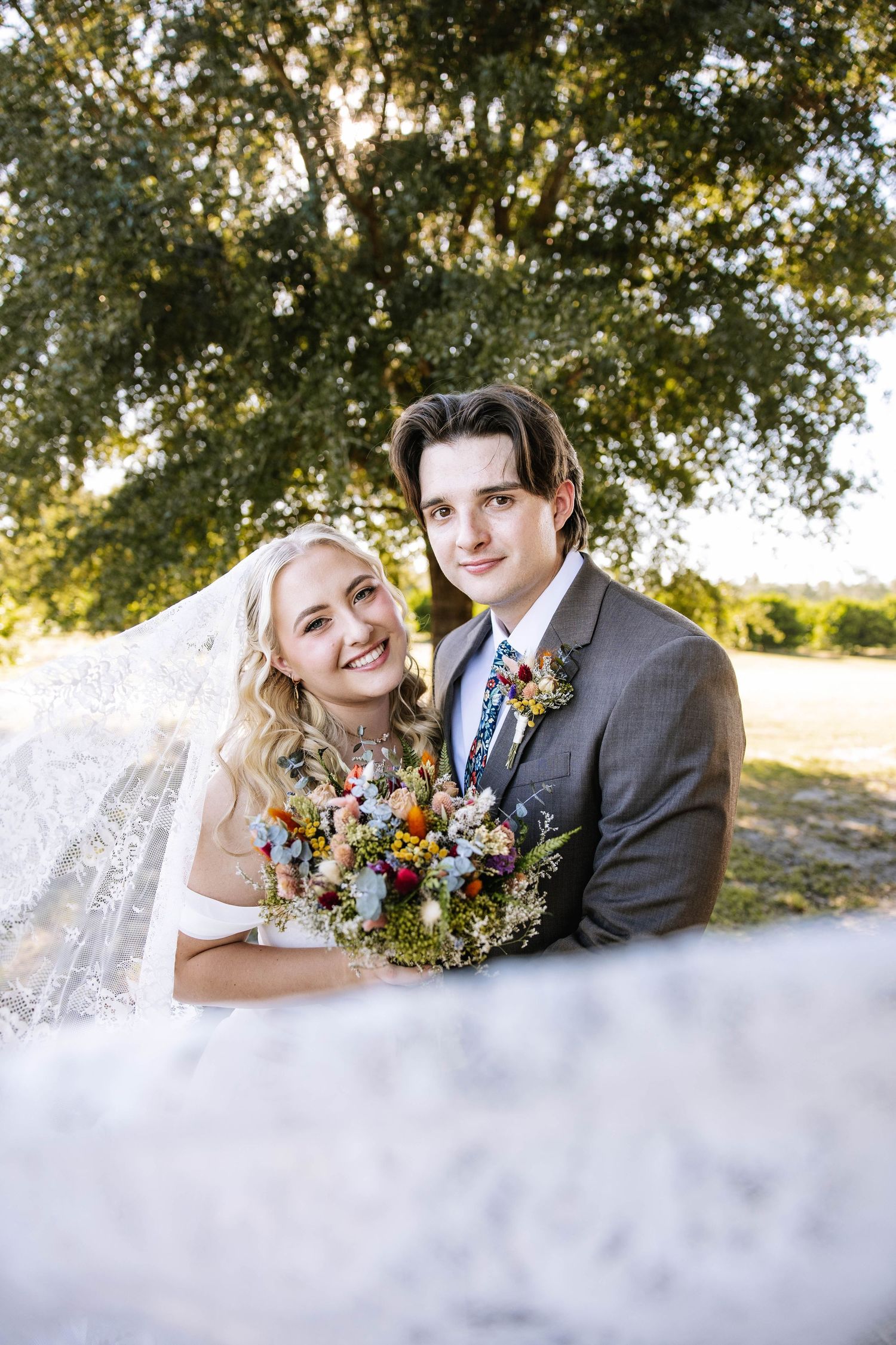 Smiling bride and groom pose outdoors under a large oak tree, bride holding colorful wildflower bouquet with lace veil flowing.