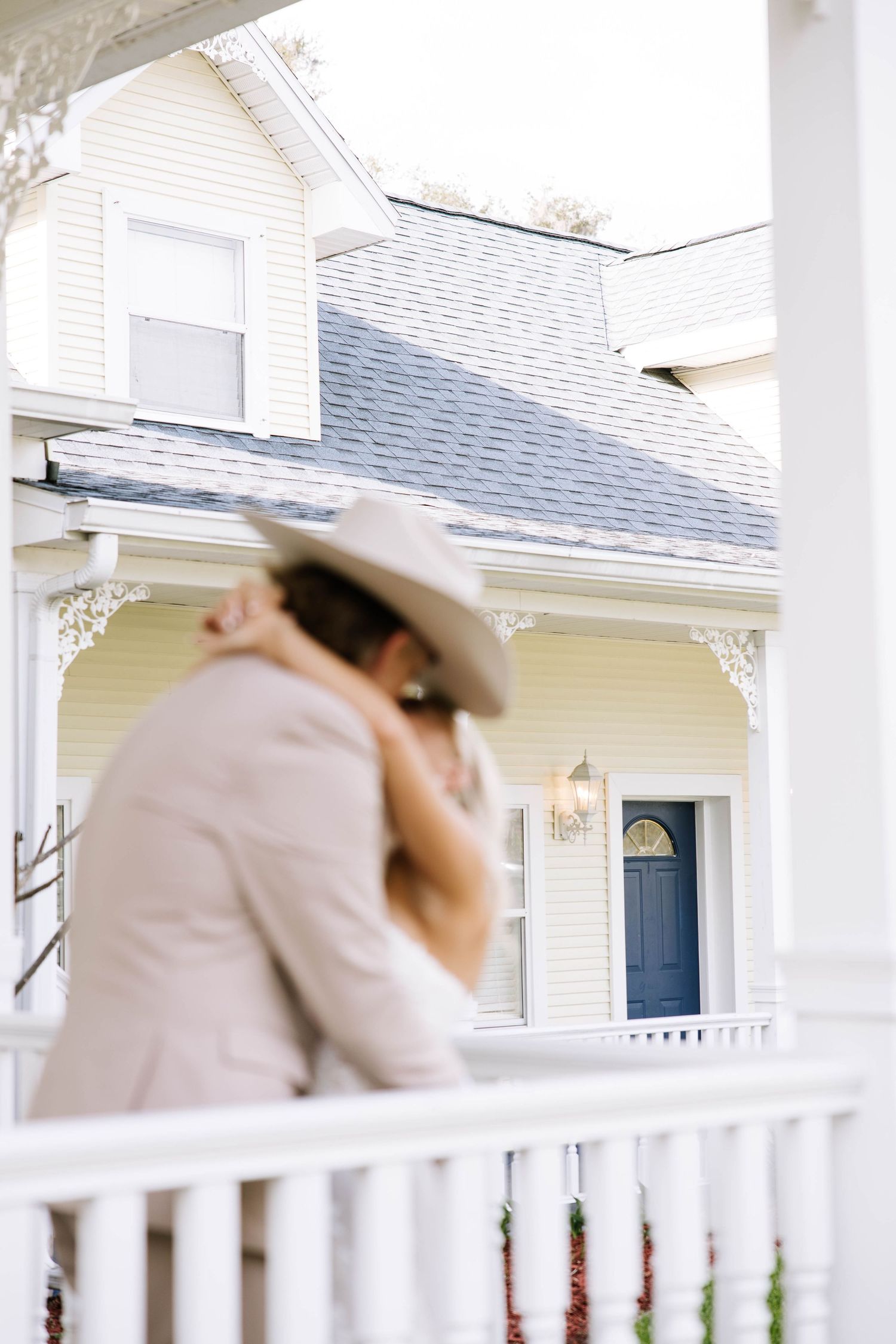 Couple sharing a romantic kiss on a white porch railing in front of a charming yellow Victorian home.