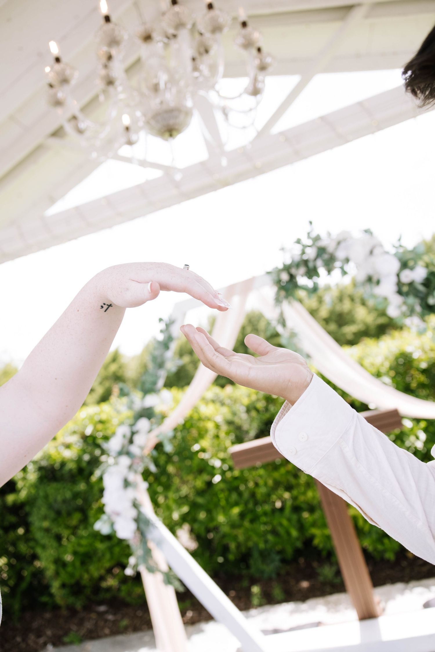 Bride and groom touching fingertips under floral chandelier at outdoor wedding ceremony.
