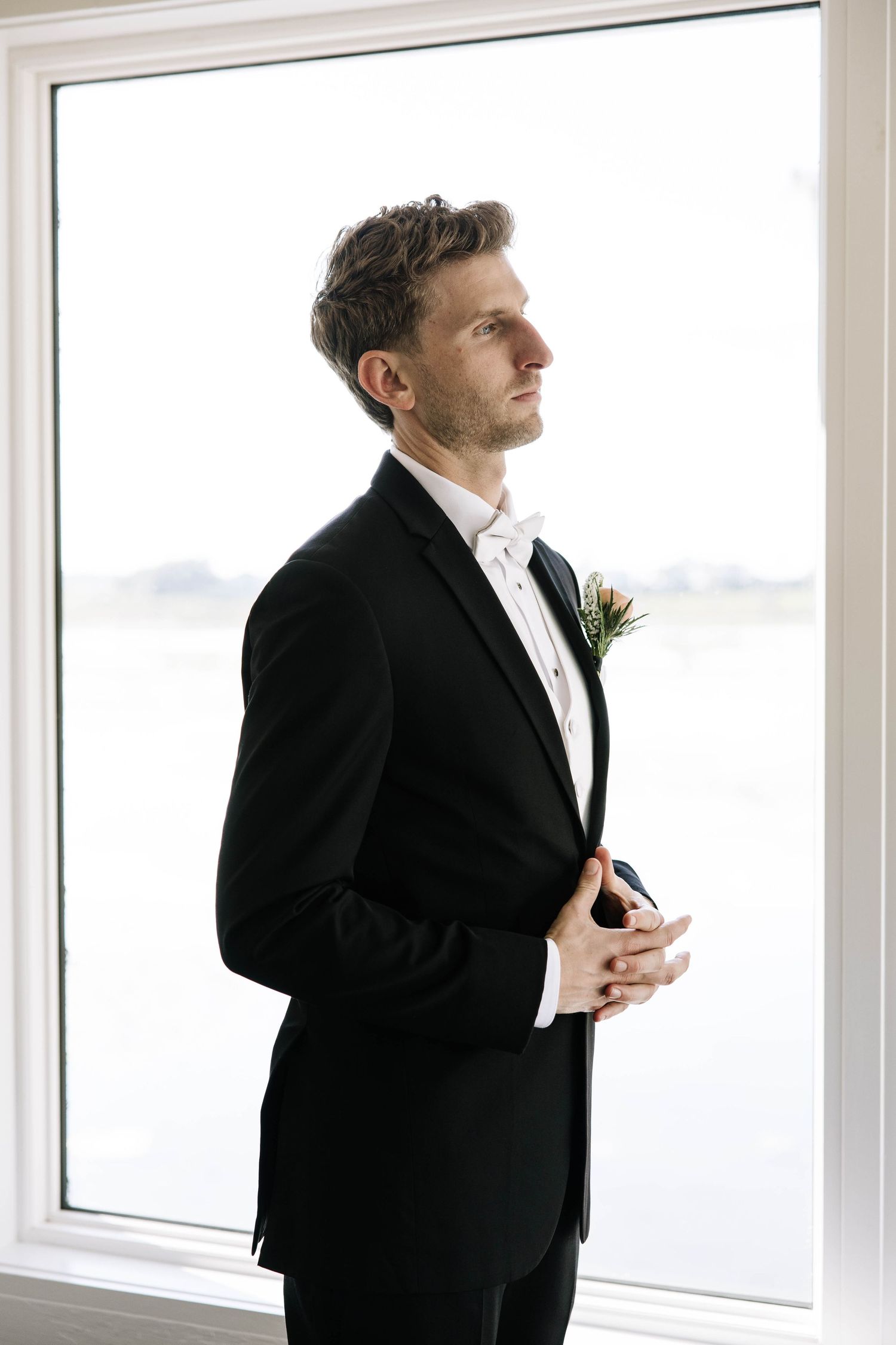 Dapper groom in black tuxedo with boutonniere gazes thoughtfully out large window with water view.