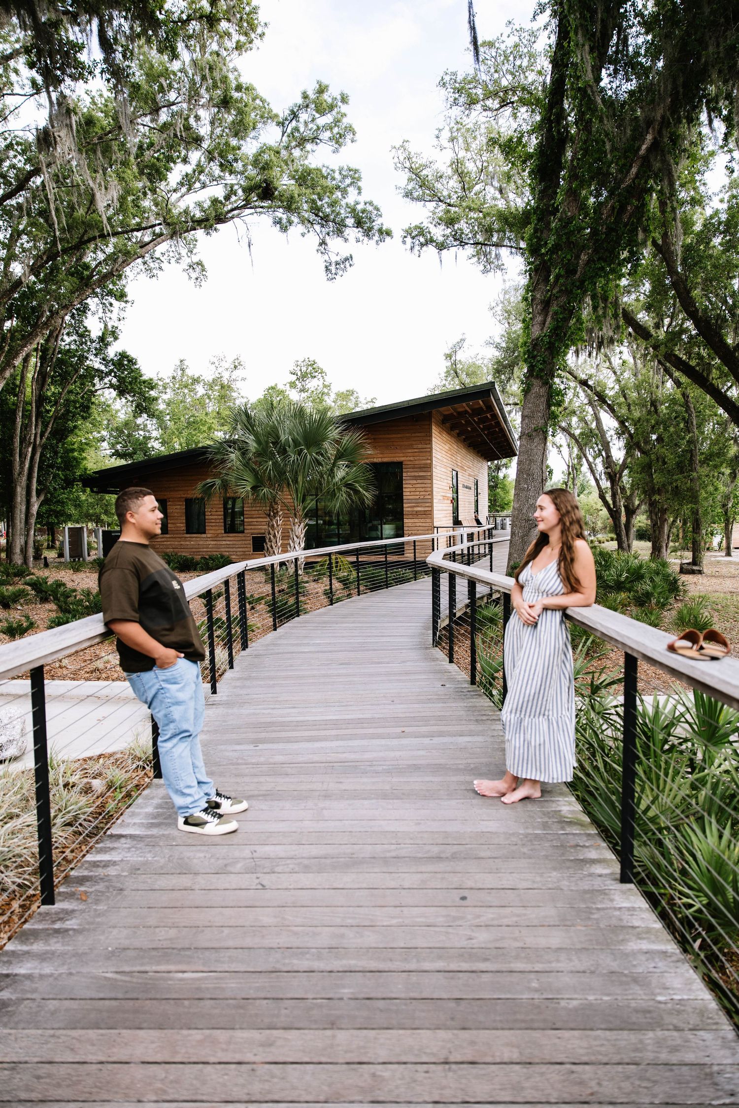 A couple stands apart on a wooden boardwalk bridge surrounded by lush trees, smiling at each other.