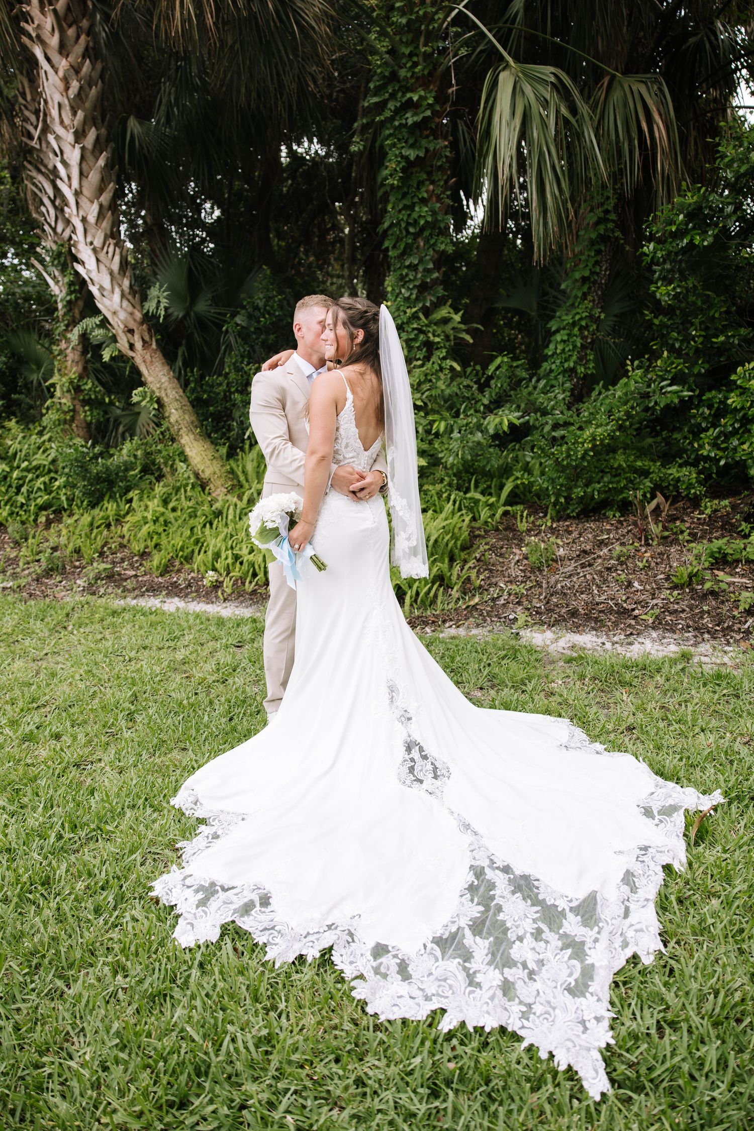 Bride and groom embrace in lush garden, showcasing bride's stunning lace cathedral train gown.