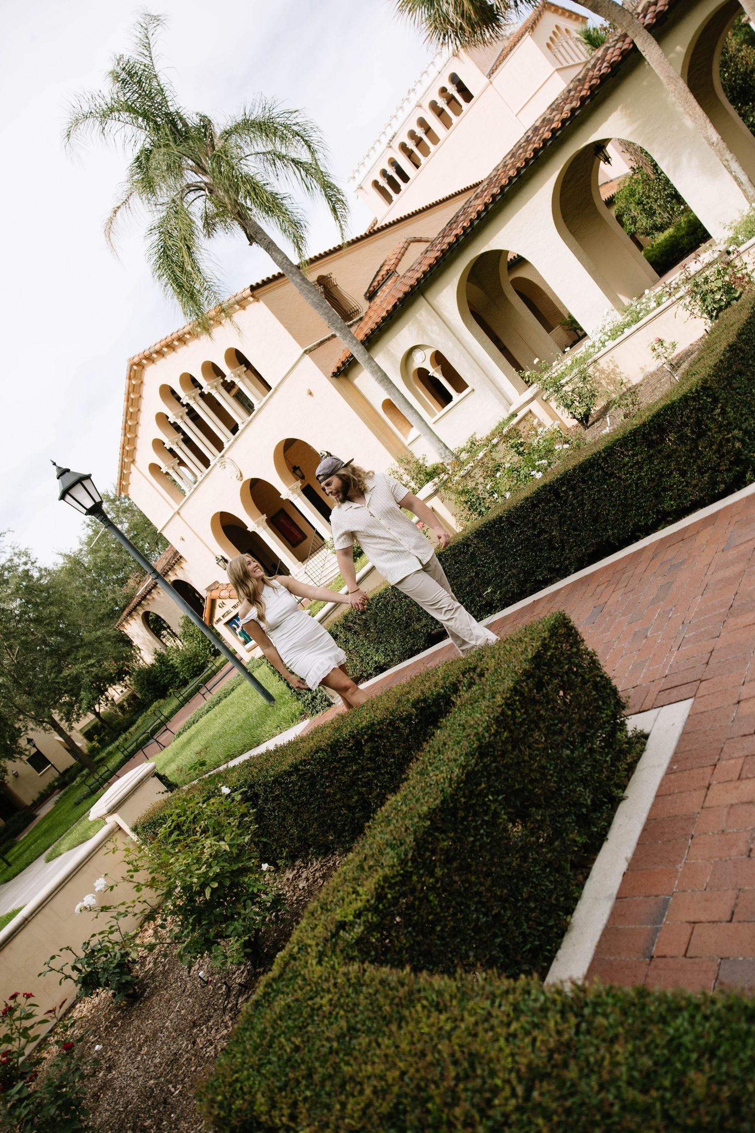 Couple walking through elegant Mediterranean-style garden with manicured hedges and arched building architecture.