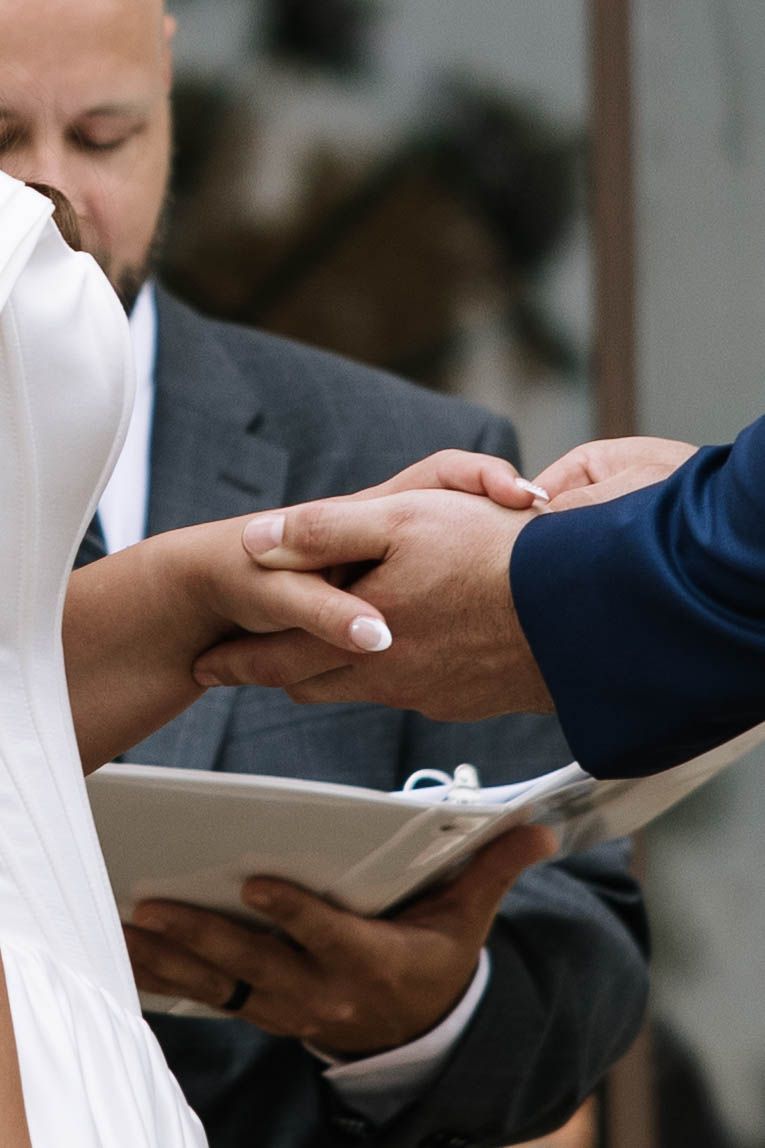 Bride and groom holding hands during wedding ring exchange ceremony with officiant holding book.