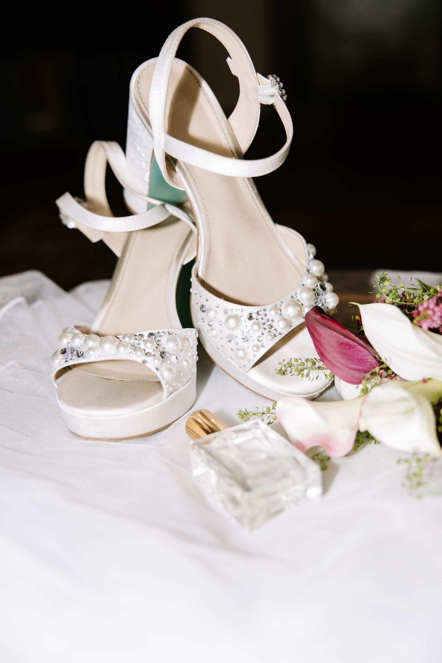 Elegant white bridal heels adorned with pearl and crystal embellishments displayed beside fresh flowers and perfume bottle.