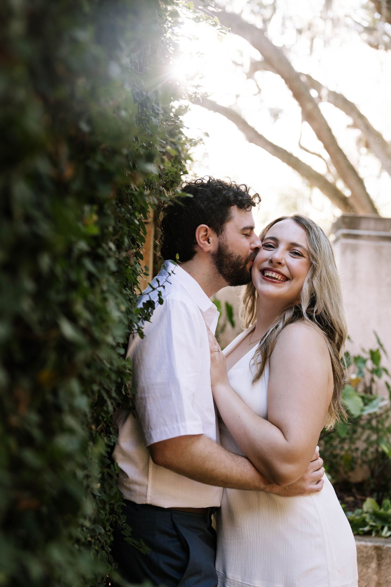 A couple embraces near an ivy-covered wall, the man kissing her cheek as she smiles radiantly in a white dress.