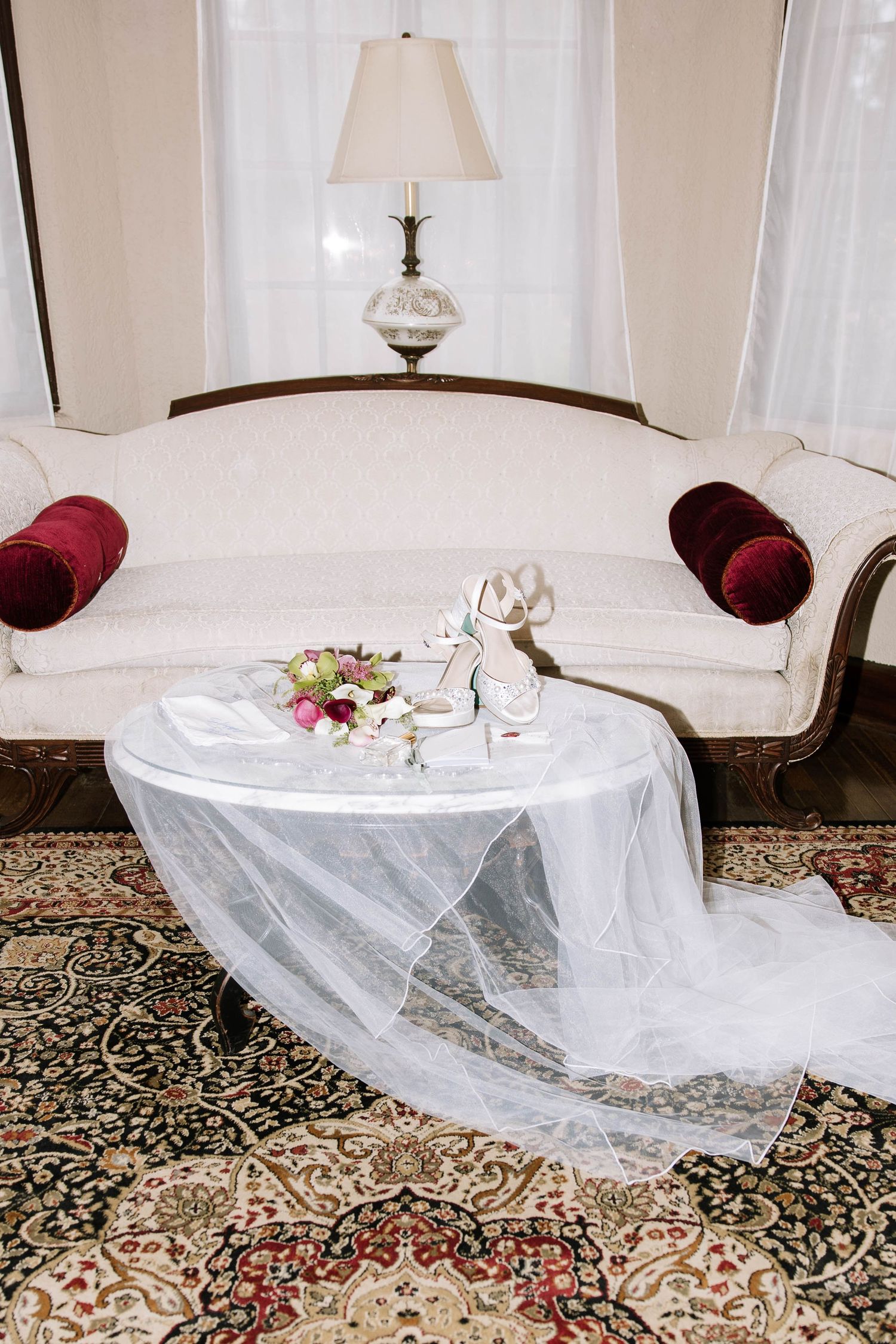 Bridal accessories including heels, a bouquet, and a flowing veil arranged on a table before an elegant sofa.